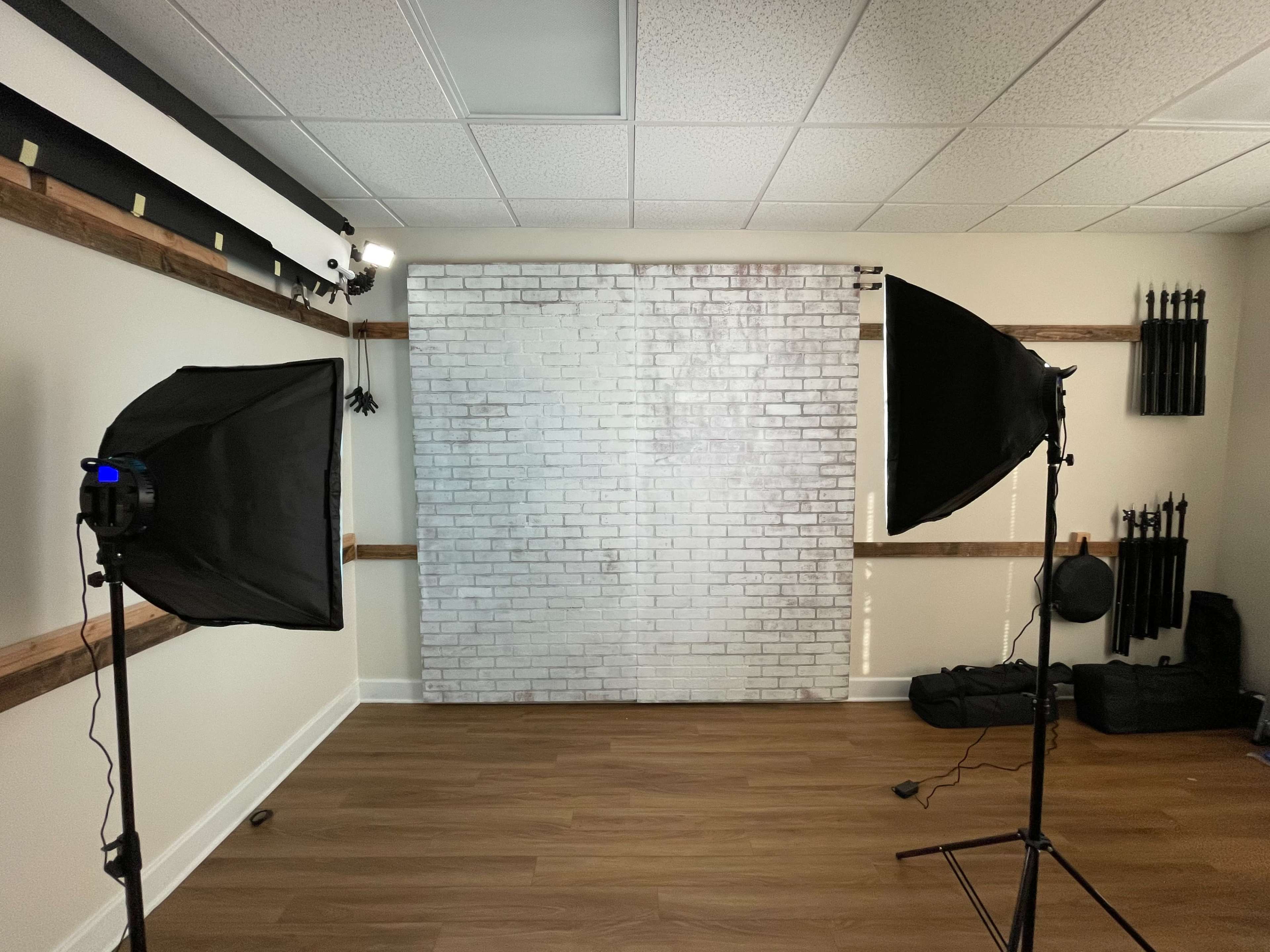 A photography studio with two softbox lights facing a white brick backdrop, surrounded by wooden accents and various equipment.