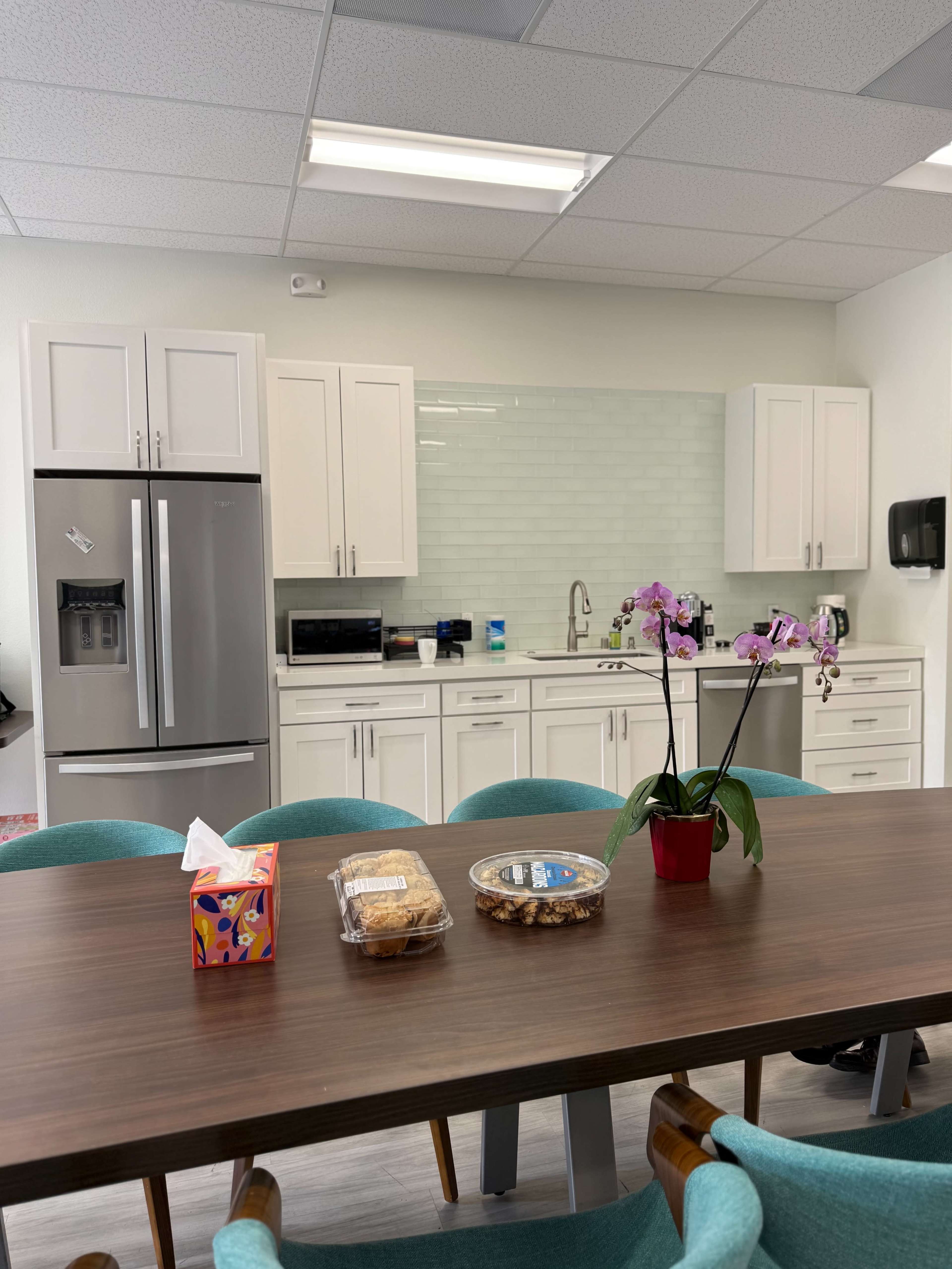 The image shows a modern kitchen and dining area with a large wooden table, equipped with a refrigerator, white cabinets, and a small potted orchid.