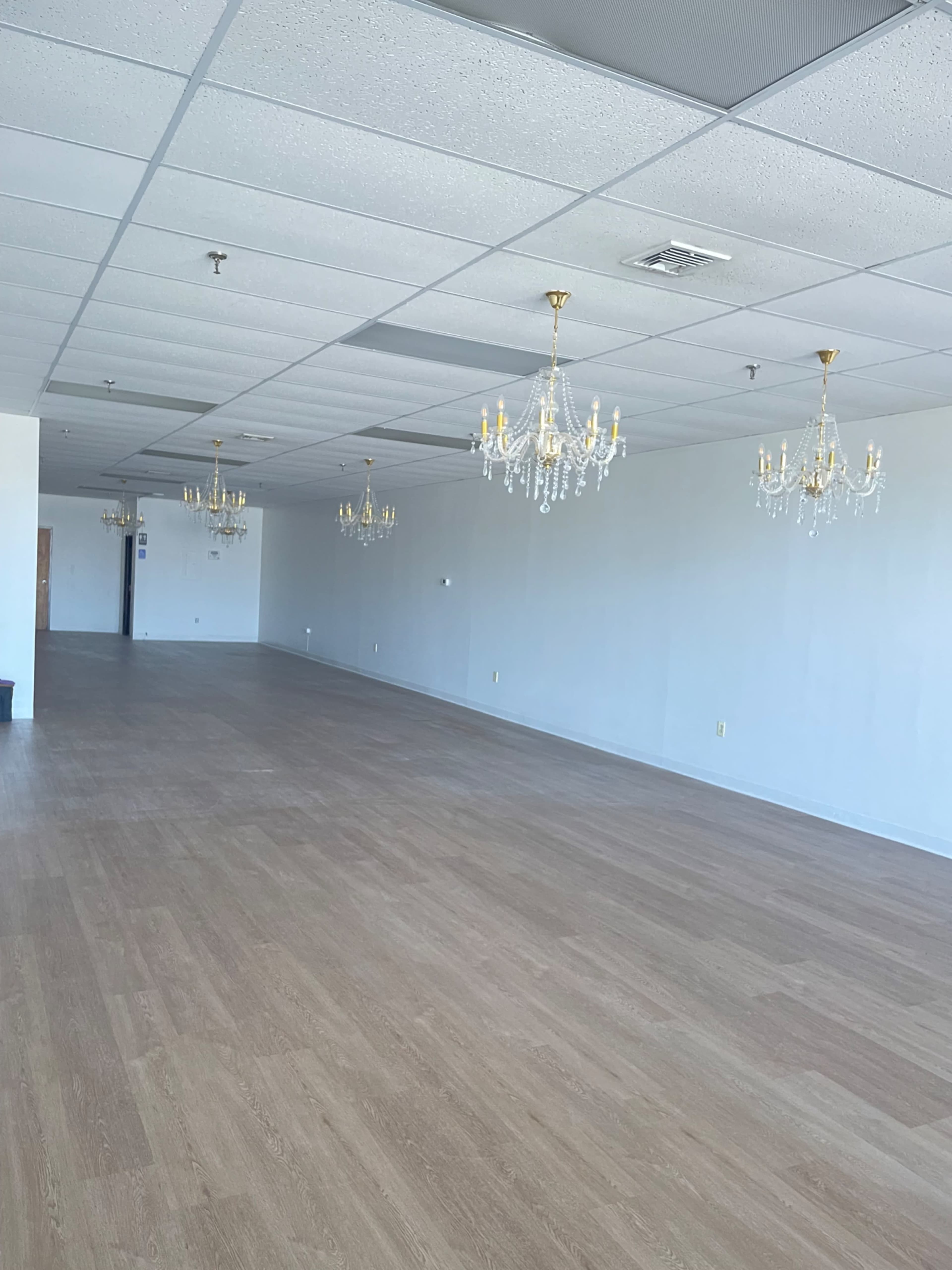 The image shows a large, empty room with light wood flooring and several chandeliers hanging from the ceiling.