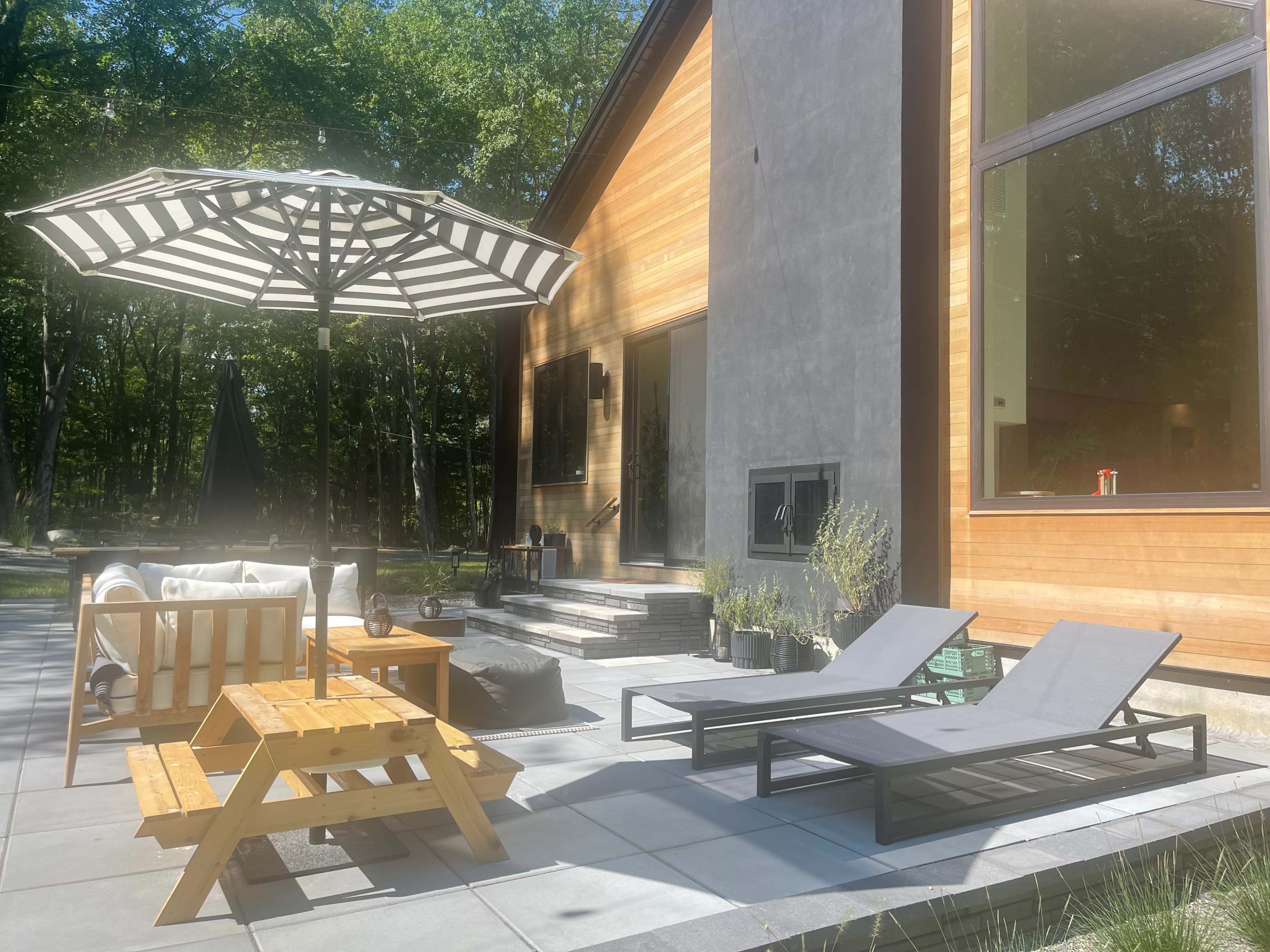 A modern outdoor patio features lounge chairs, a picnic table, and a large striped umbrella next to a house with a wooden and concrete facade.