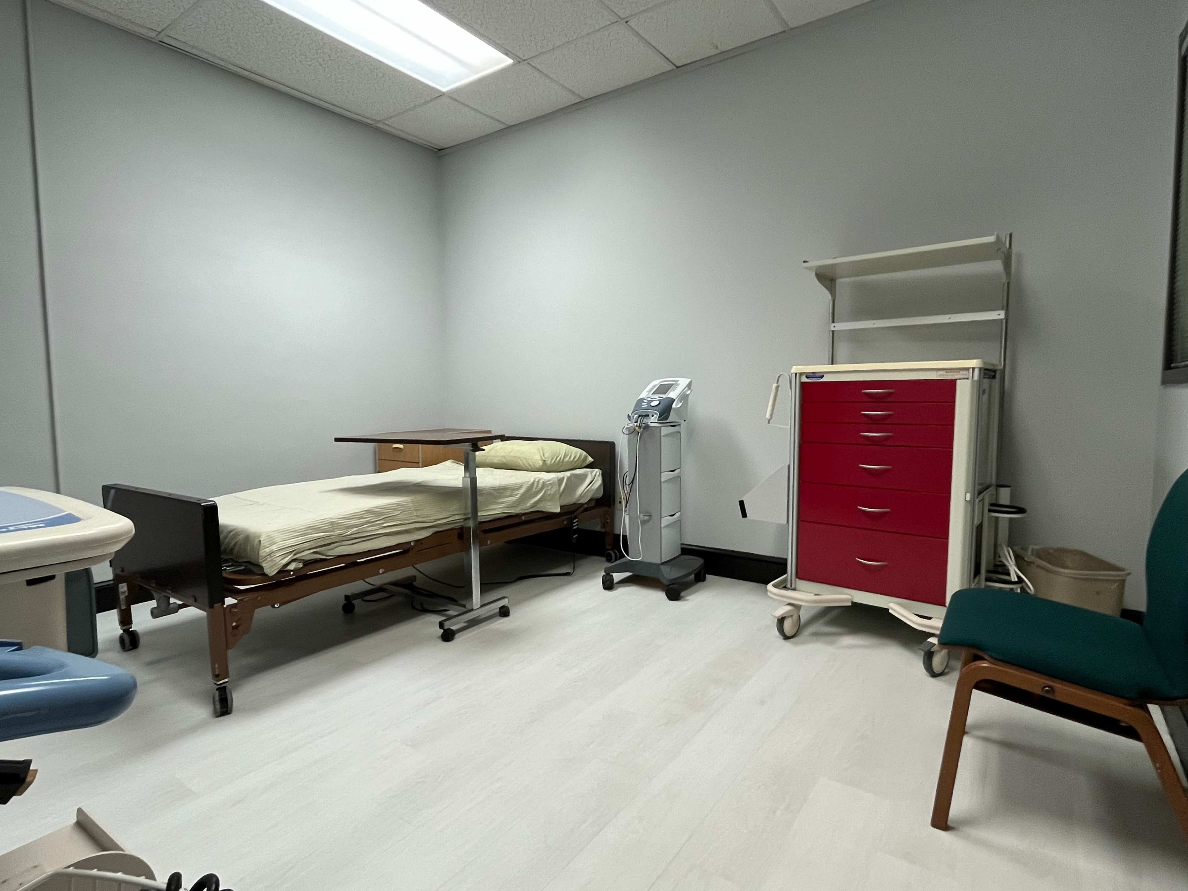 The image shows a medical examination room with a bed, a medical equipment stand, a mobile cart with drawers, and a chair.