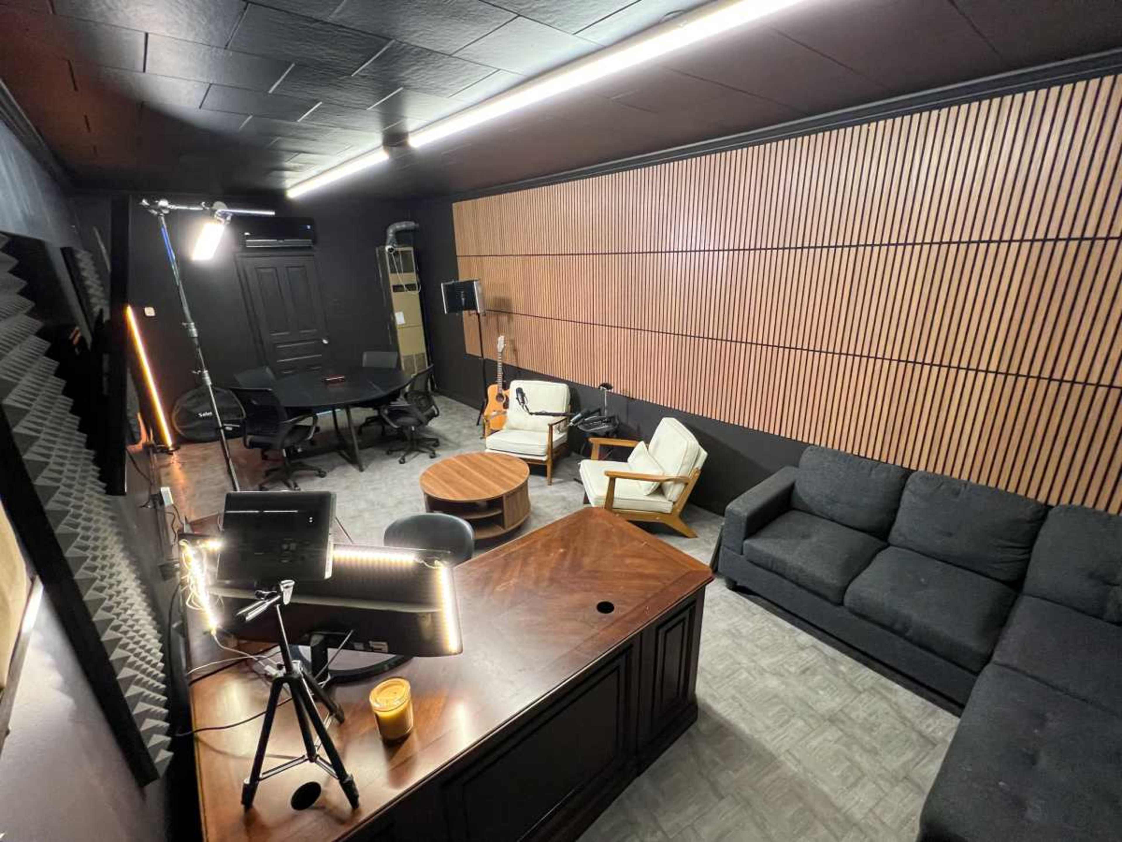 The image shows a dimly lit room with acoustic paneling, featuring a dark sectional sofa, a round coffee table, a guitar, and a workspace with a monitor and camera setup.