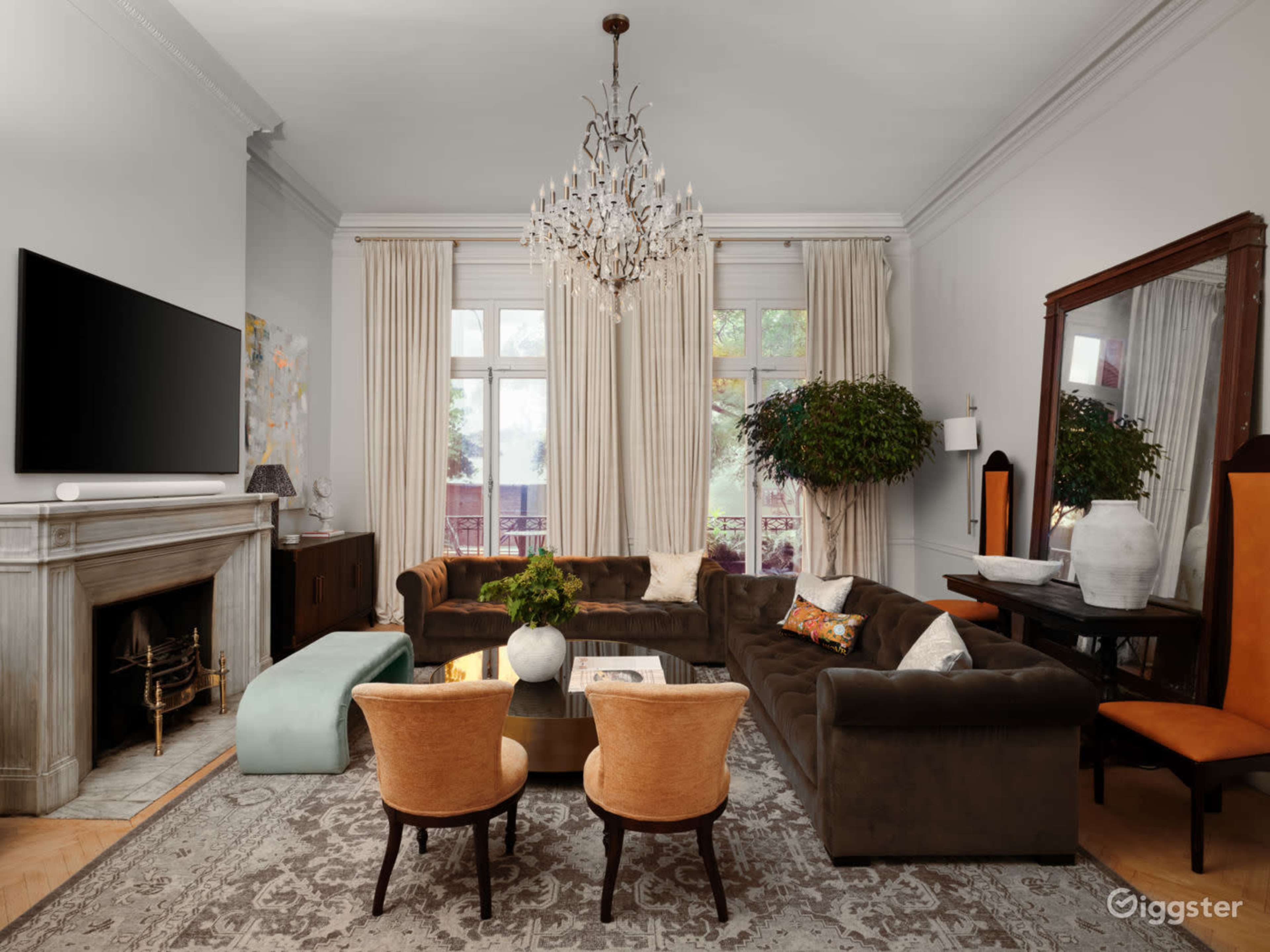 The living room features a large, dark sofa arranged around a central coffee table, with a chandelier hanging above and large windows revealing greenery outside.