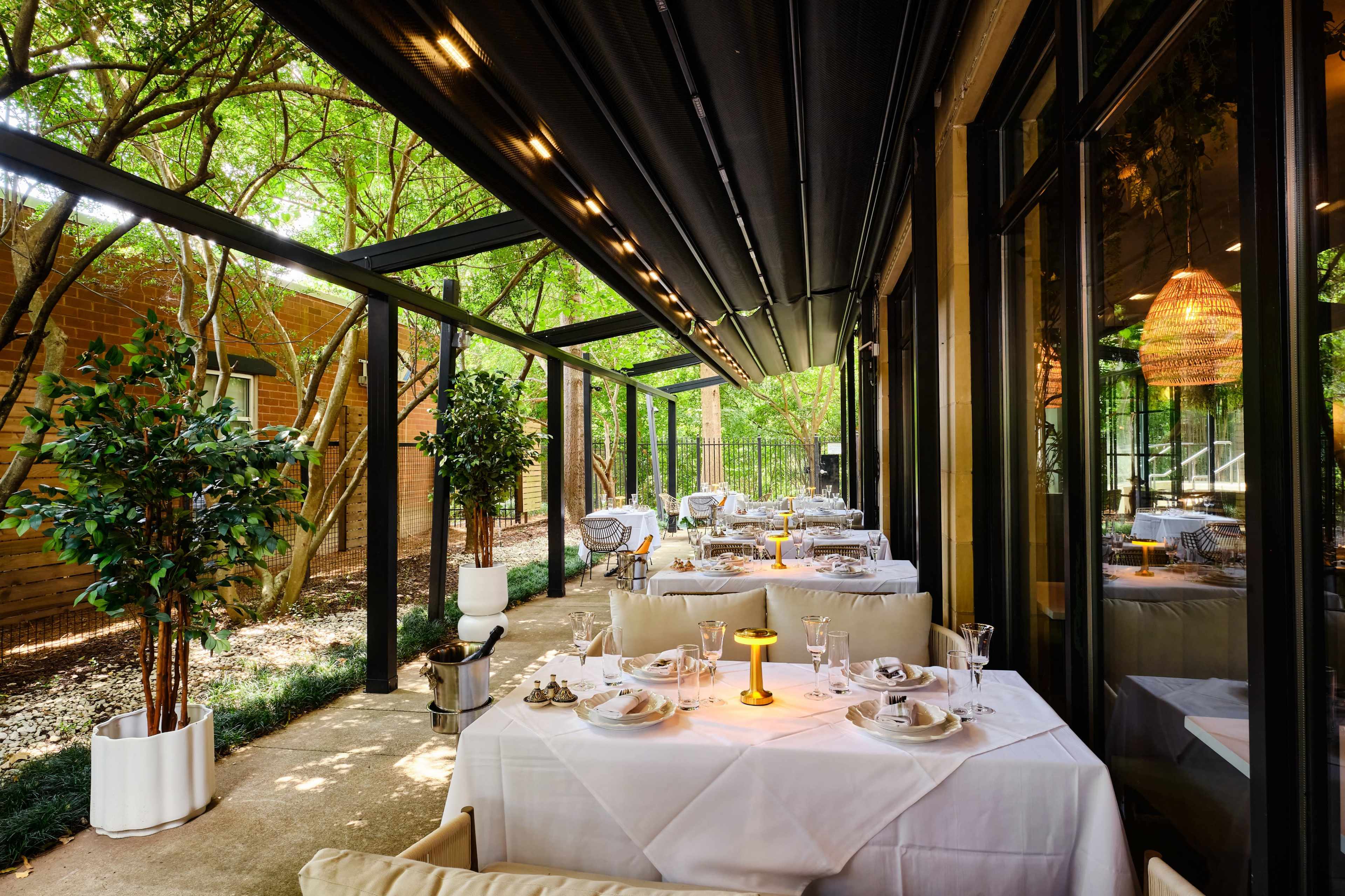 The image shows a modern outdoor dining area with neatly set tables, surrounded by greenery and glass walls.