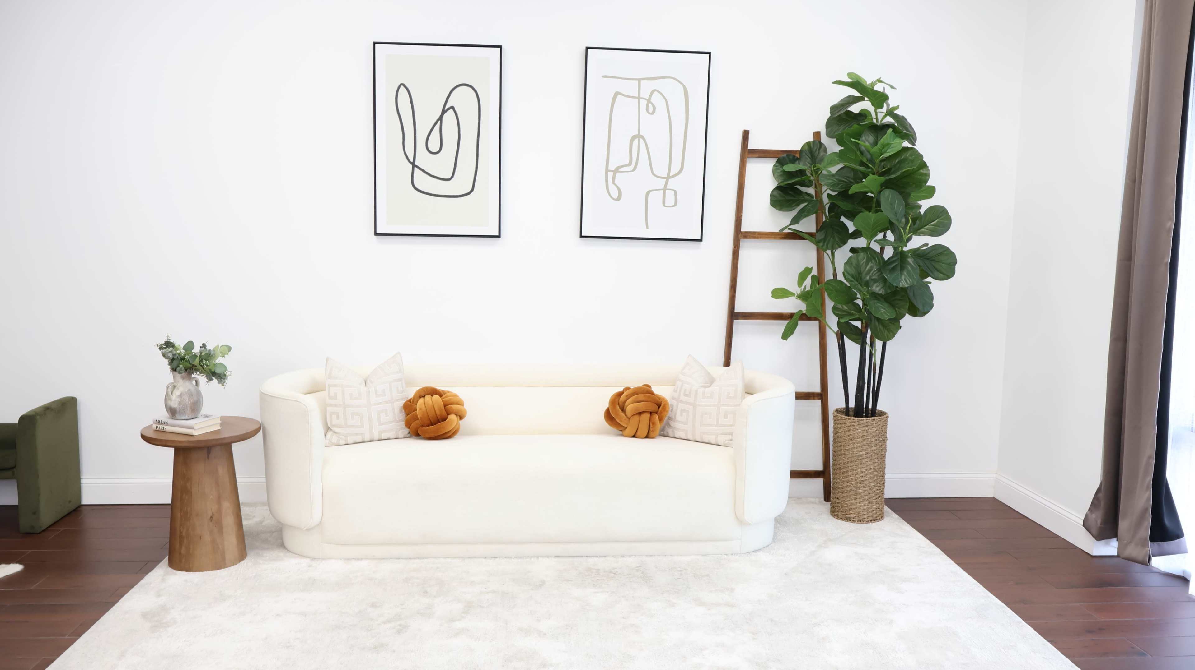 The scene features a minimalist living room with a white sofa adorned with decorative pillows, two framed line art prints on the wall, a tall indoor plant, and a wooden side table.