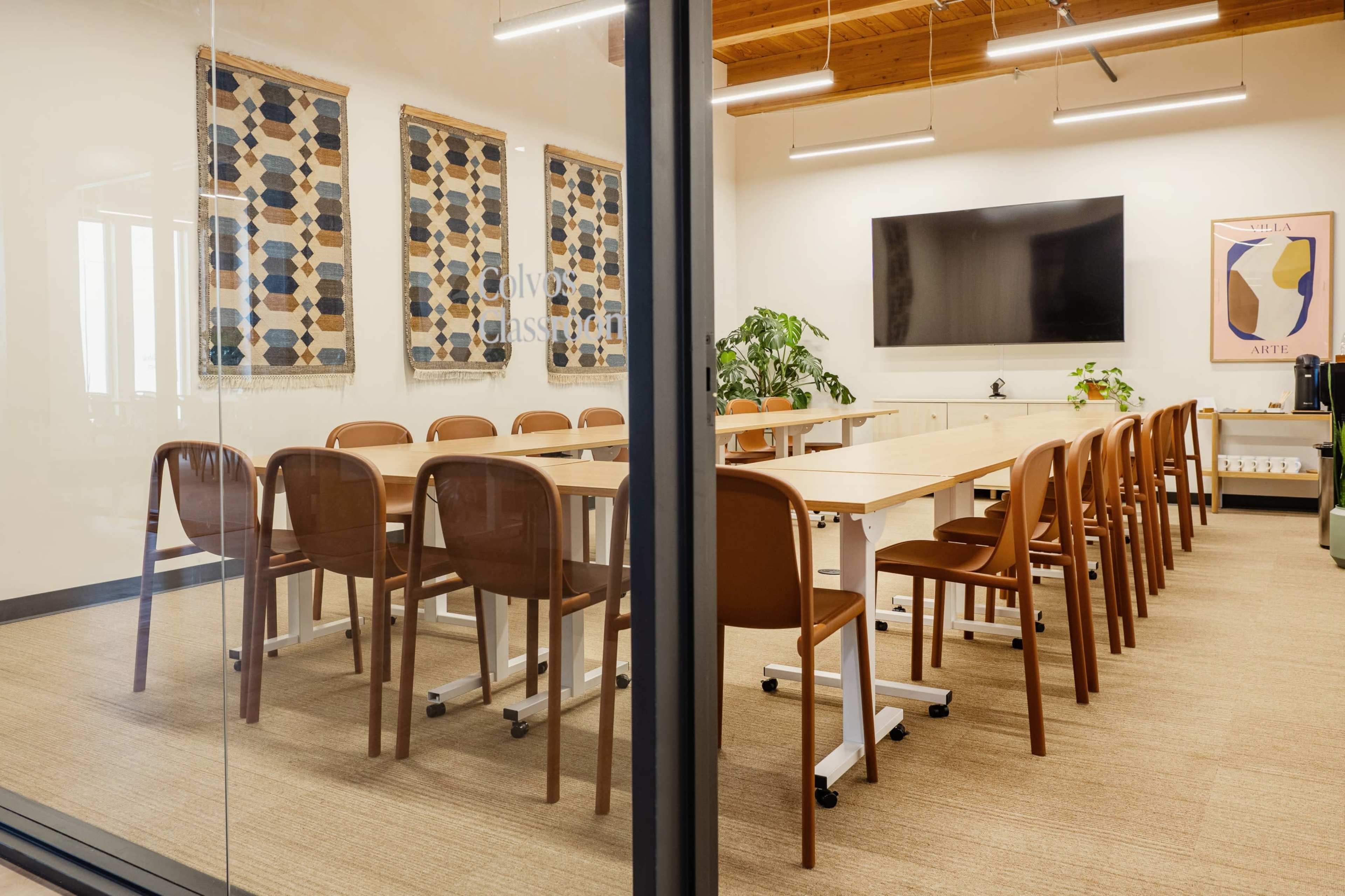 A modern classroom features wooden tables arranged in a U-shape, with brown chairs and decorative wall hangings, alongside a large screen and potted plants.