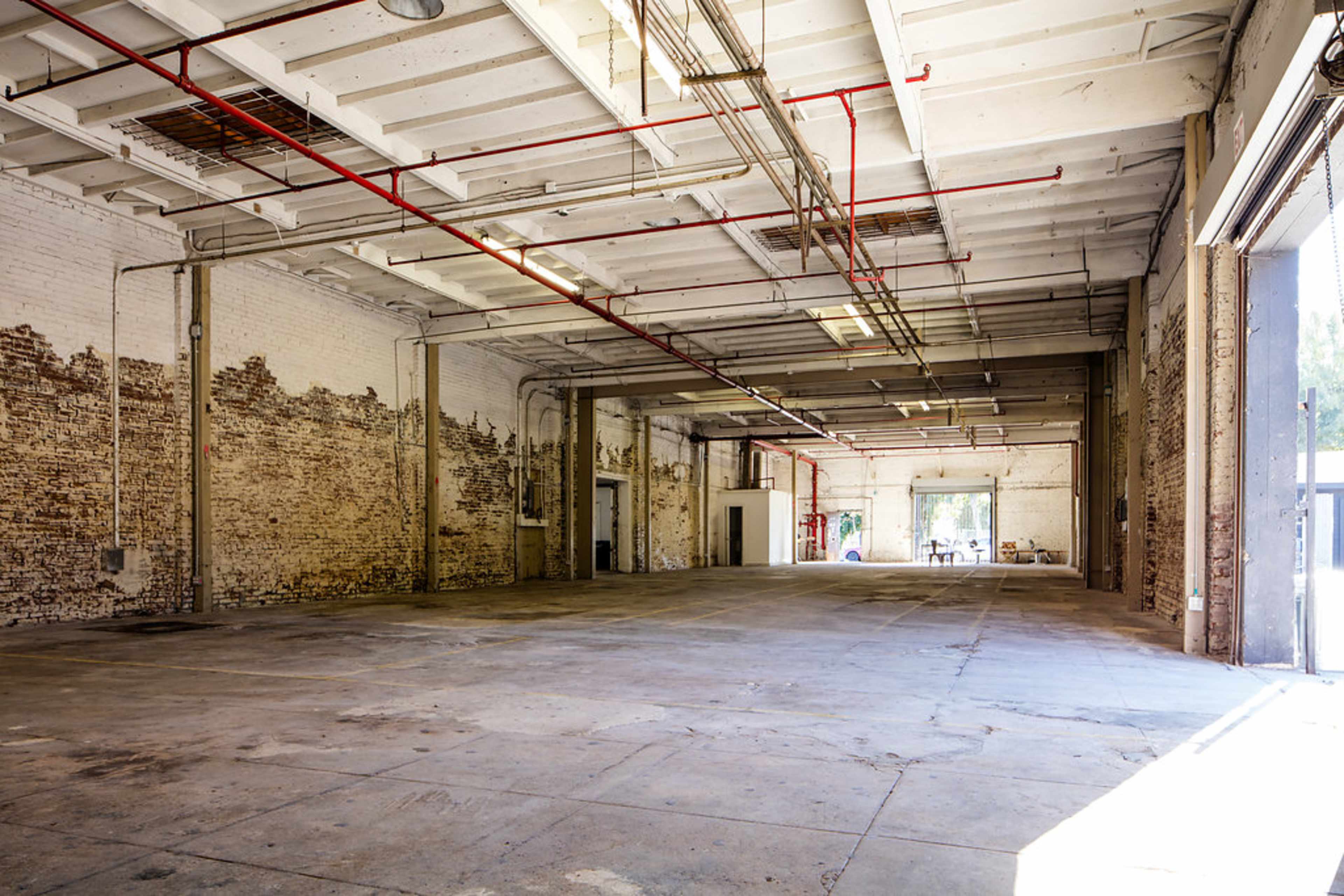 An empty, spacious warehouse with exposed brick walls and concrete flooring, featuring large windows that allow natural light to enter.