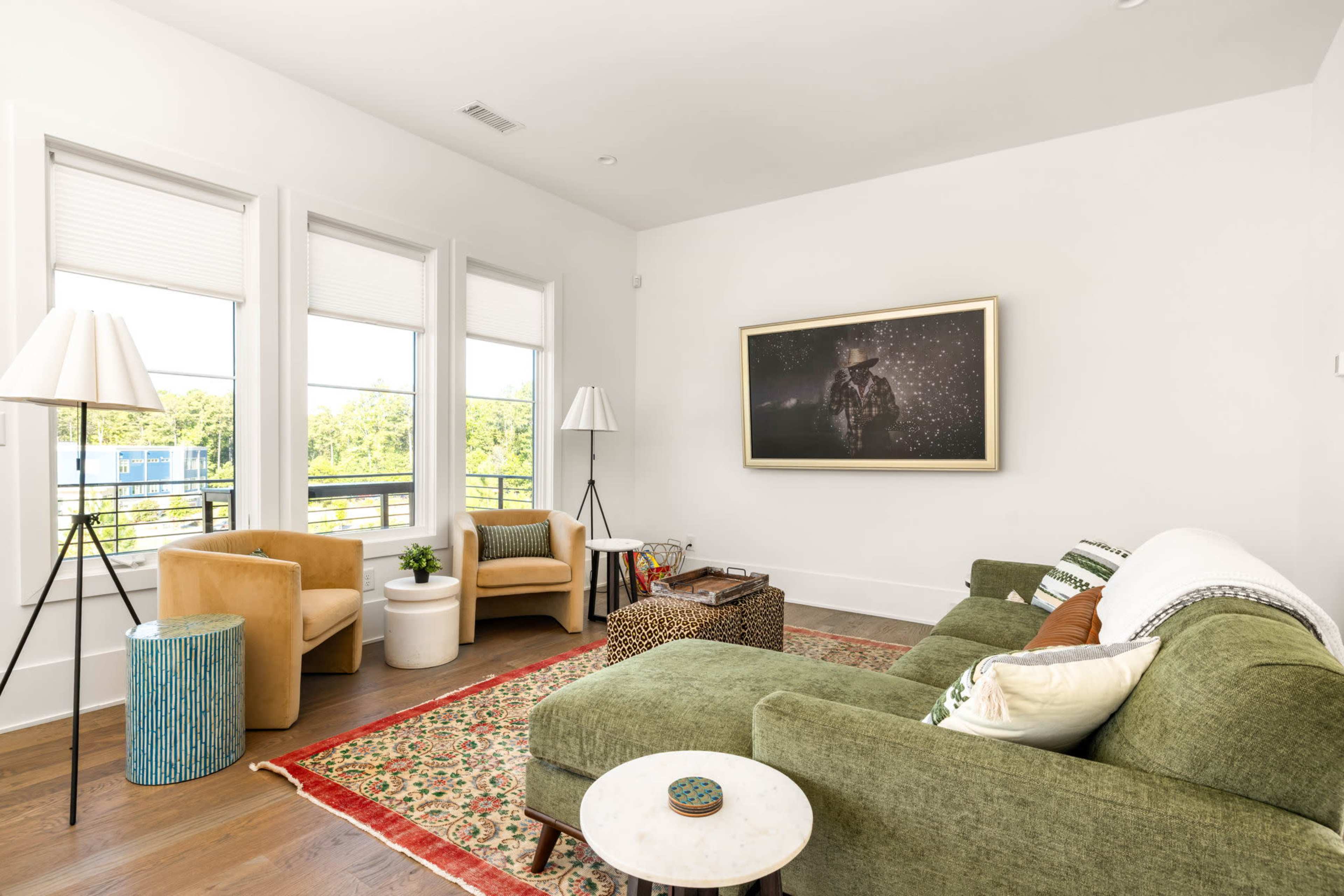 The image shows a modern living room featuring a green sectional sofa, two beige armchairs, a round coffee table, and large windows with shades, complemented by a rug on the hardwood floor.