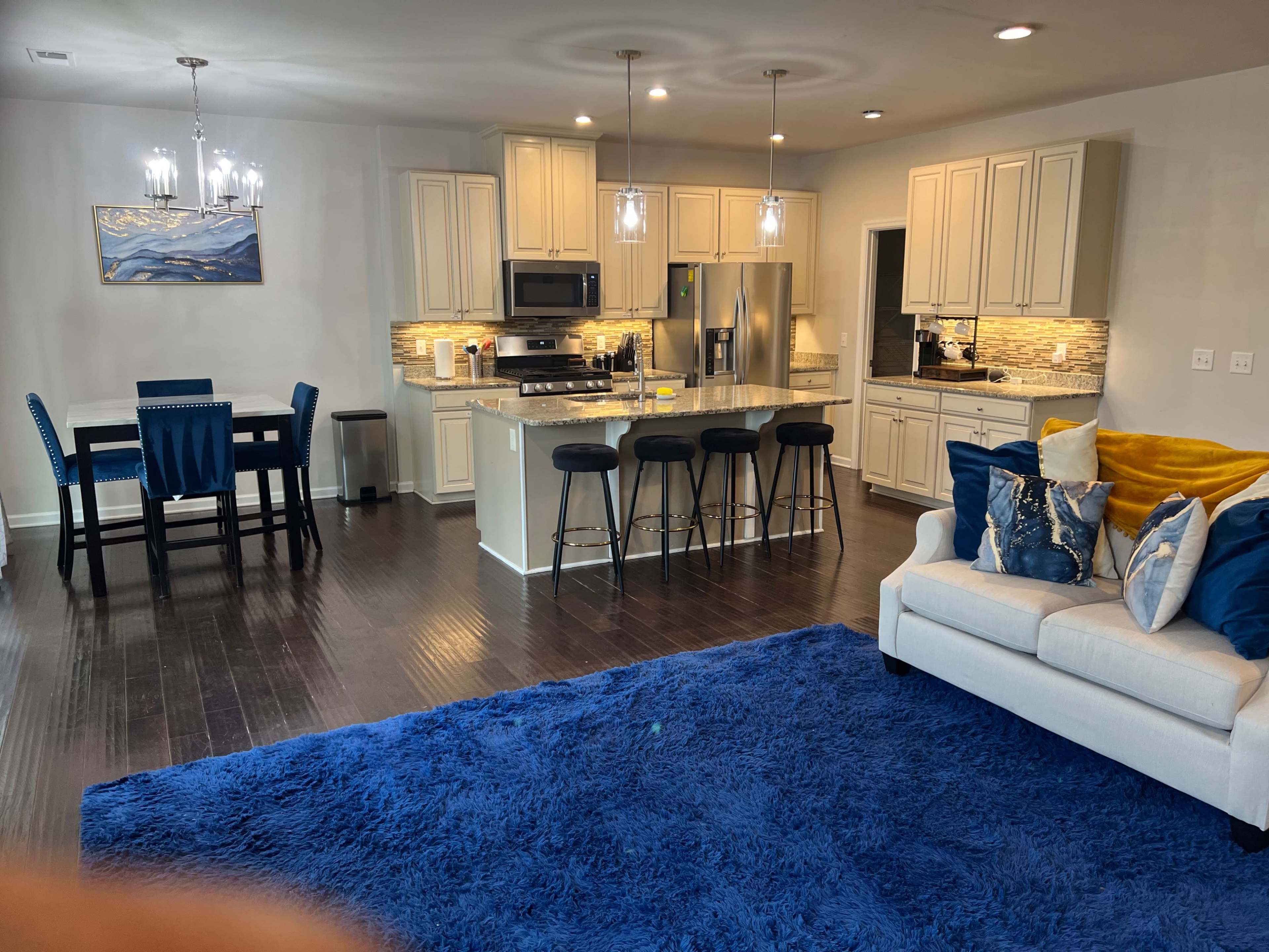 A modern open-concept kitchen and living area with a blue area rug, a white sofa, a dining table, and bar stools at the kitchen island.
