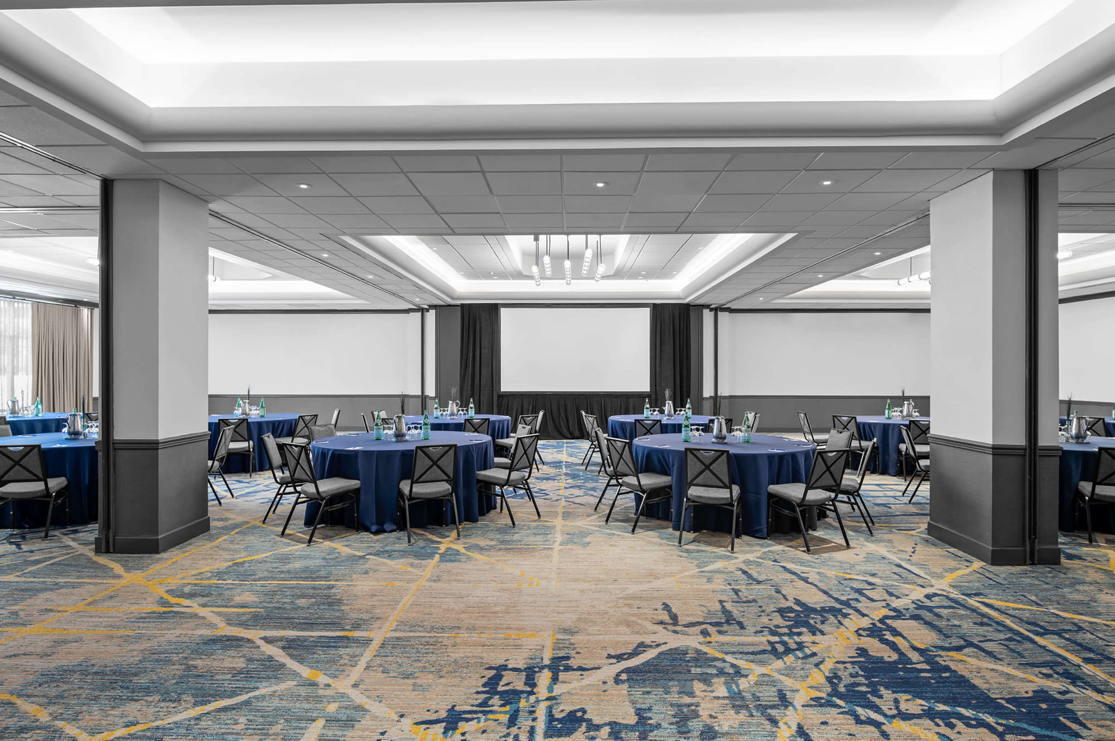A spacious event room features multiple round tables dressed in blue tablecloths, surrounded by black chairs, with a large projection screen at the front.