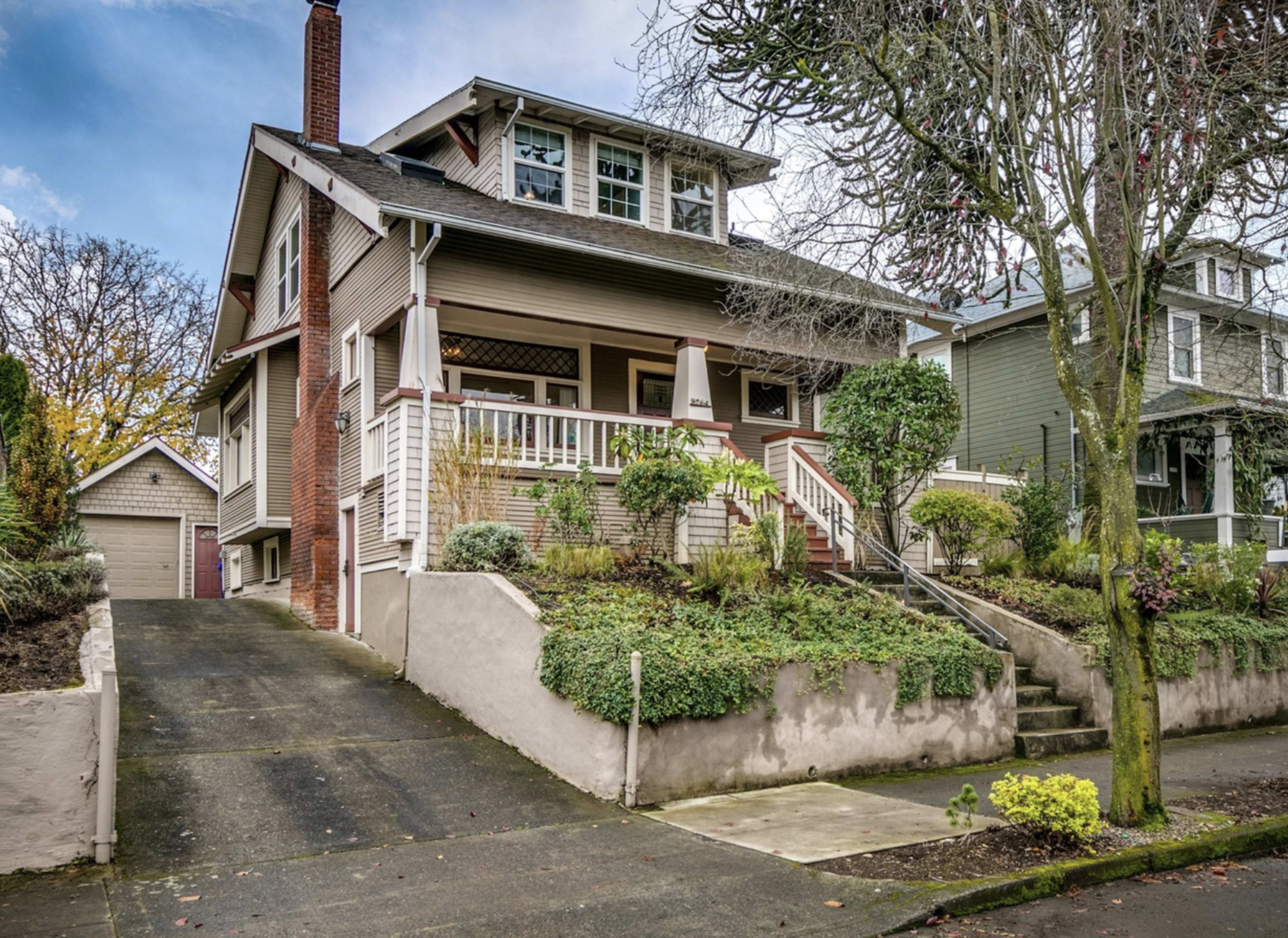 A large, two-story home with a front porch is situated on a sloped driveway, surrounded by landscaped greenery and trees.
