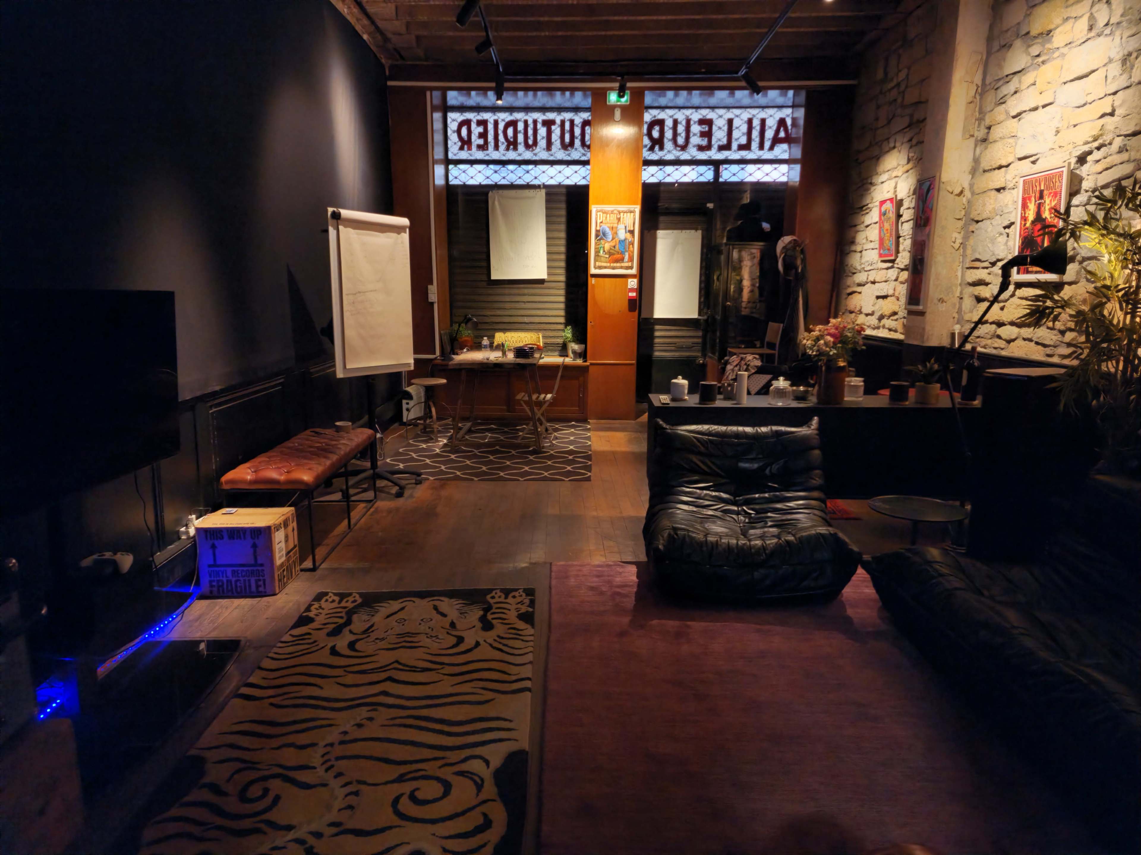 The image shows a dimly lit lounge area with a large black beanbag, a patterned rug, a couch, and a presentation board in a room featuring exposed brick walls and a window with text.