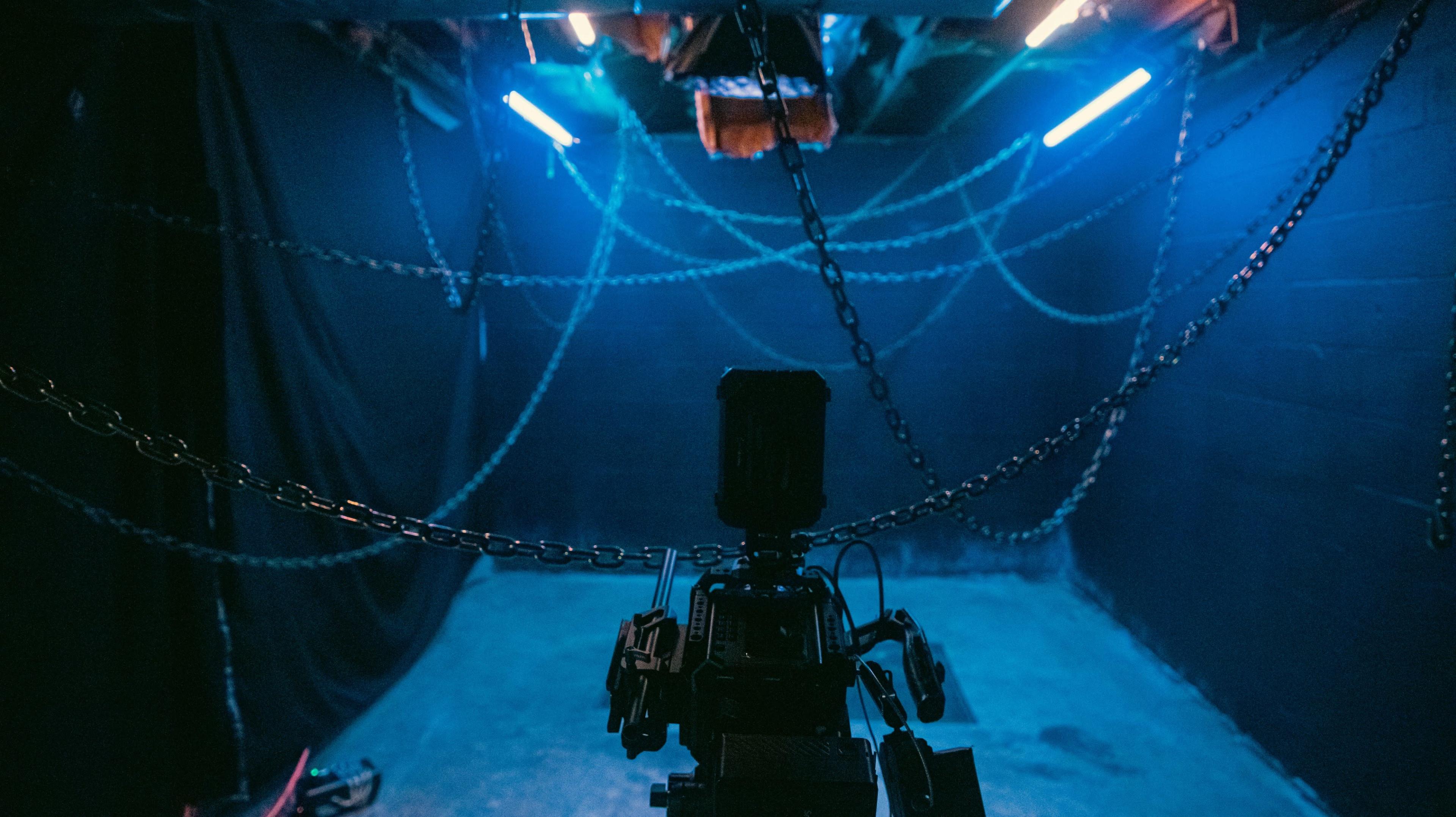 A camera is positioned in a dimly lit room with chains hanging from the ceiling and blue lights illuminating the space.