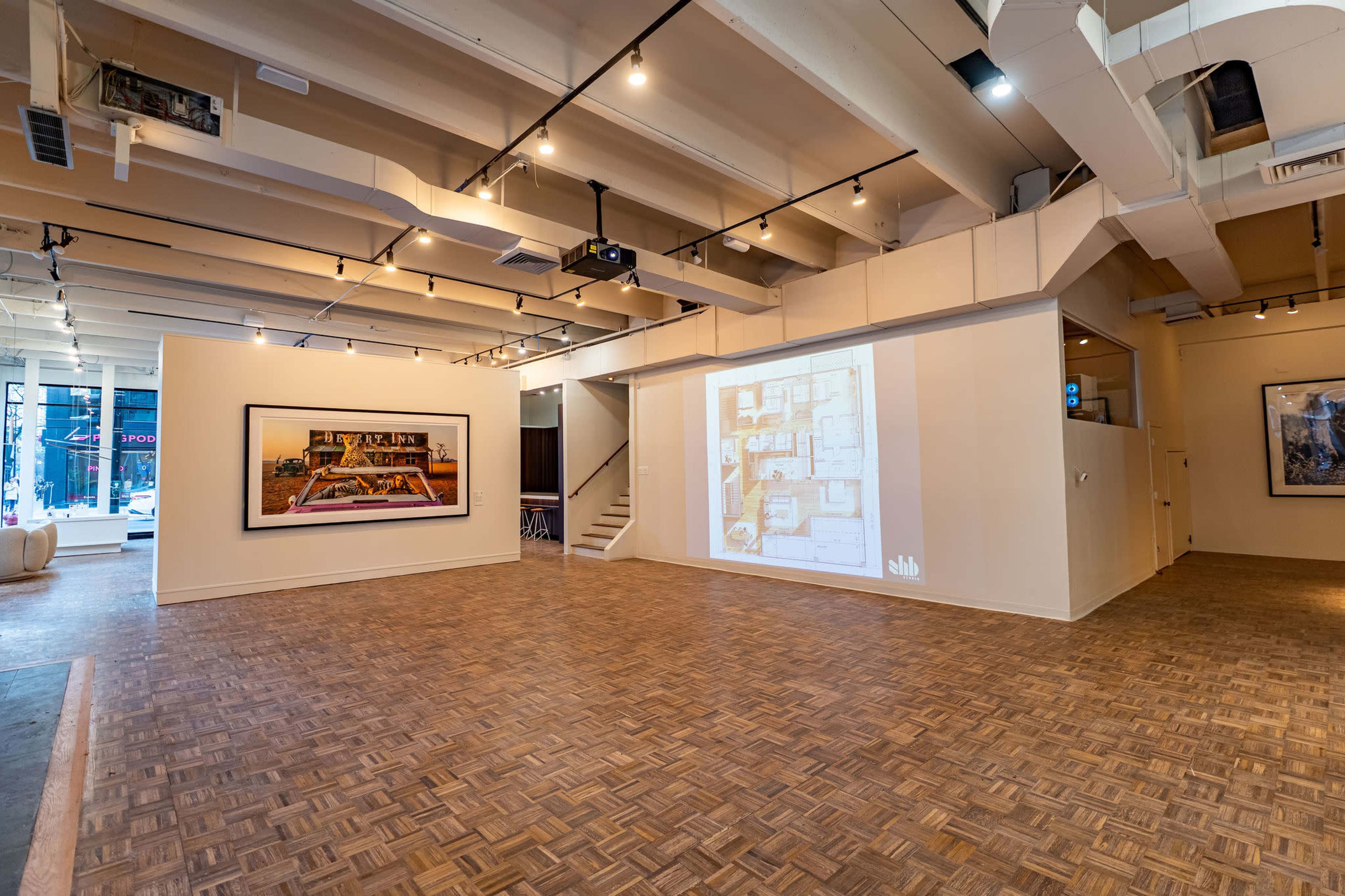 The image shows a spacious art gallery featuring wooden flooring, bright lighting, and framed artwork displayed on the walls.