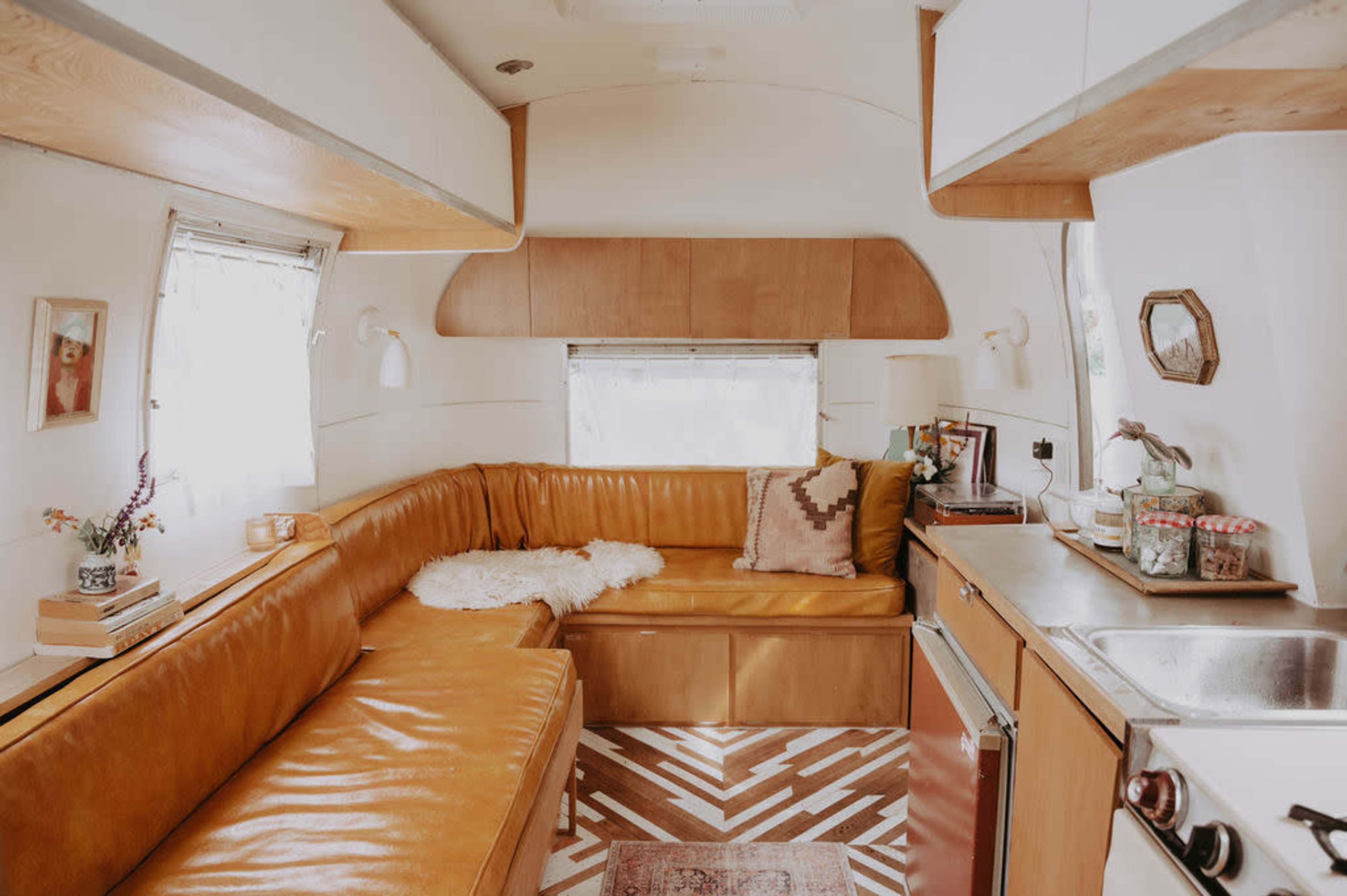 The interior of a vintage travel trailer features a cozy seating area with orange leather cushions, a small kitchen with a sink, and decorative elements on the walls.