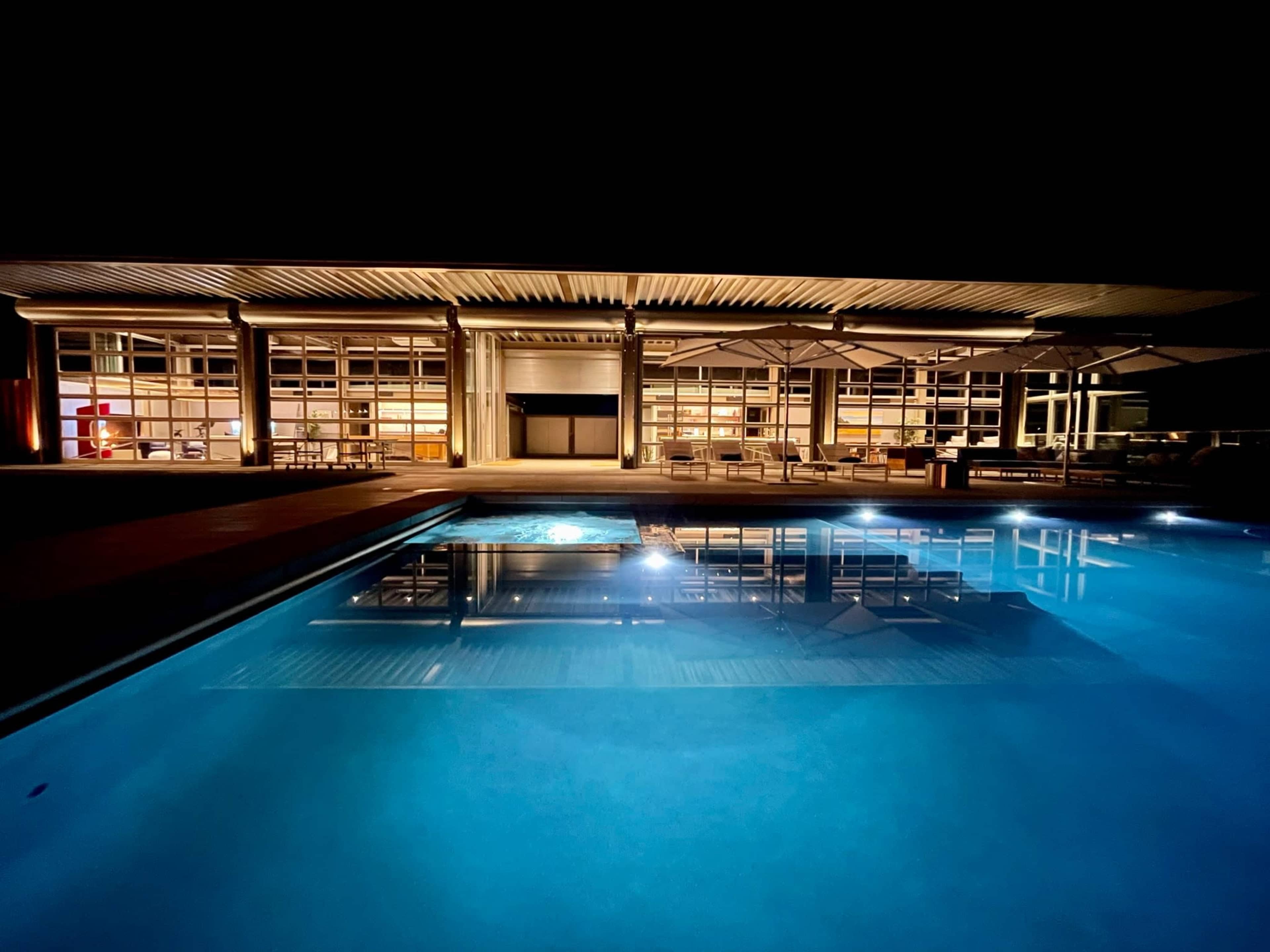 A modern house with large glass windows reflects in a well-lit swimming pool at night.