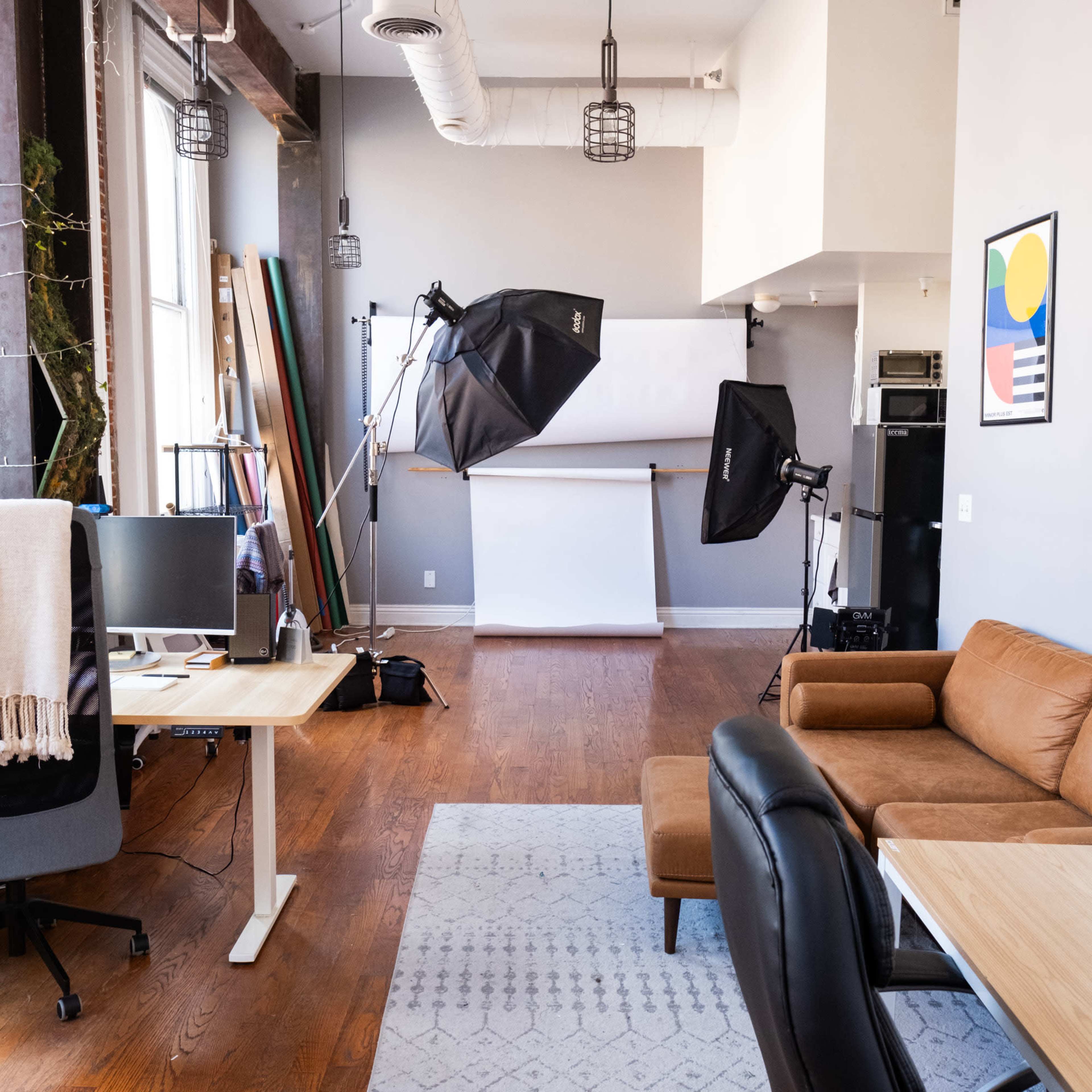 The image shows a spacious studio workspace featuring a brown sofa, a desk with a computer, lighting equipment, and a white backdrop setup.