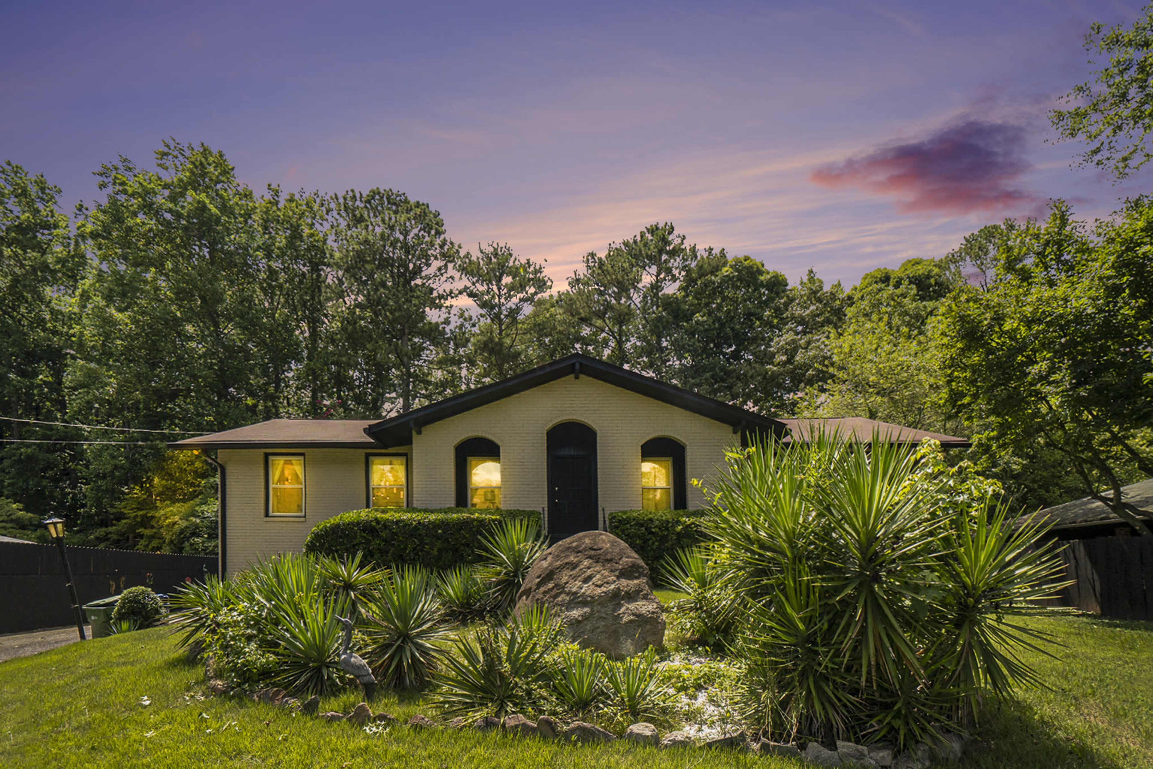 A single-story house with illuminated windows is set among landscaped greenery and trees at dusk.