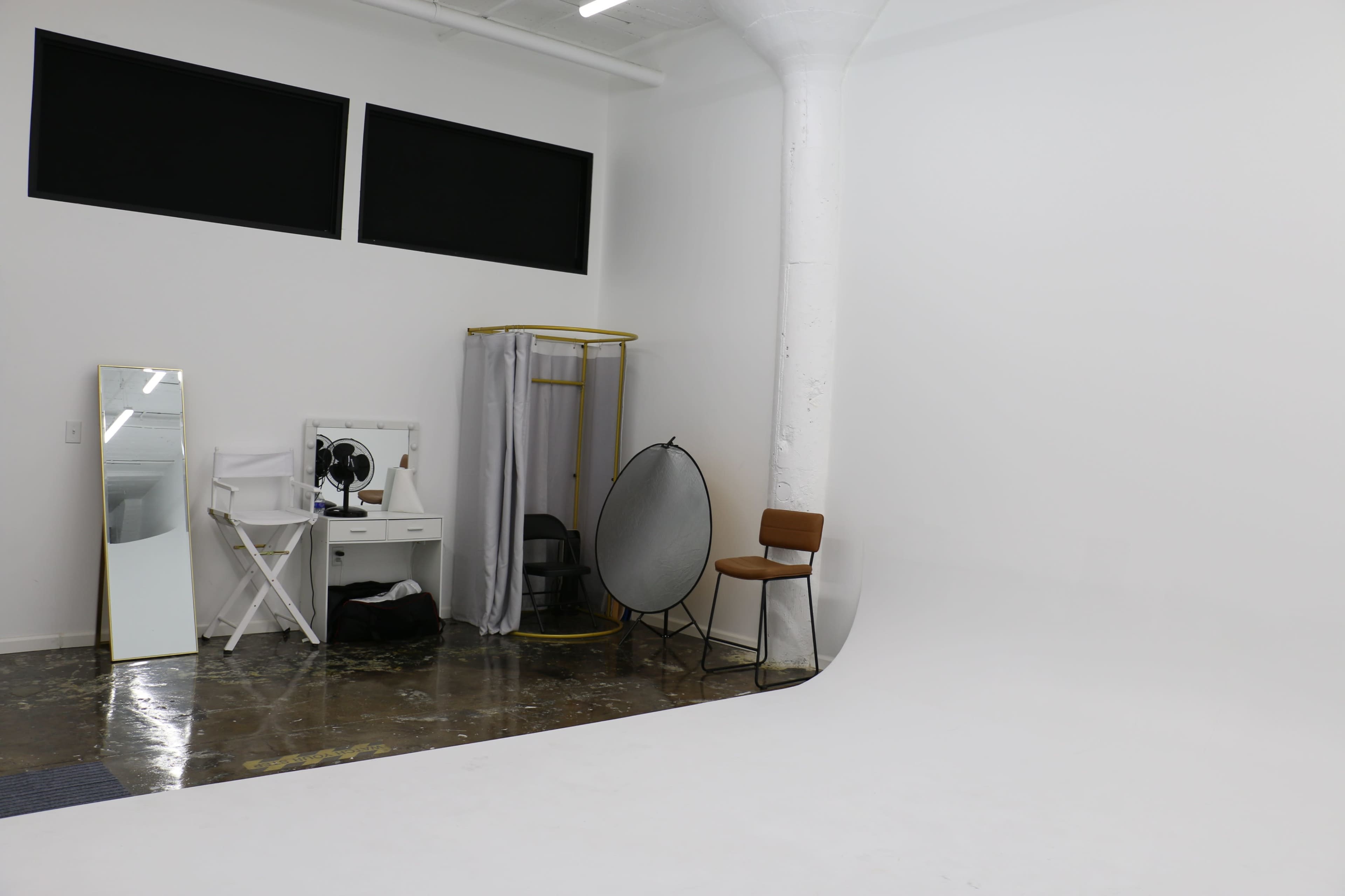 The image shows a minimalistic photography studio featuring a seamless white backdrop, a folding chair, a mirror, and various equipment for photo shoots.
