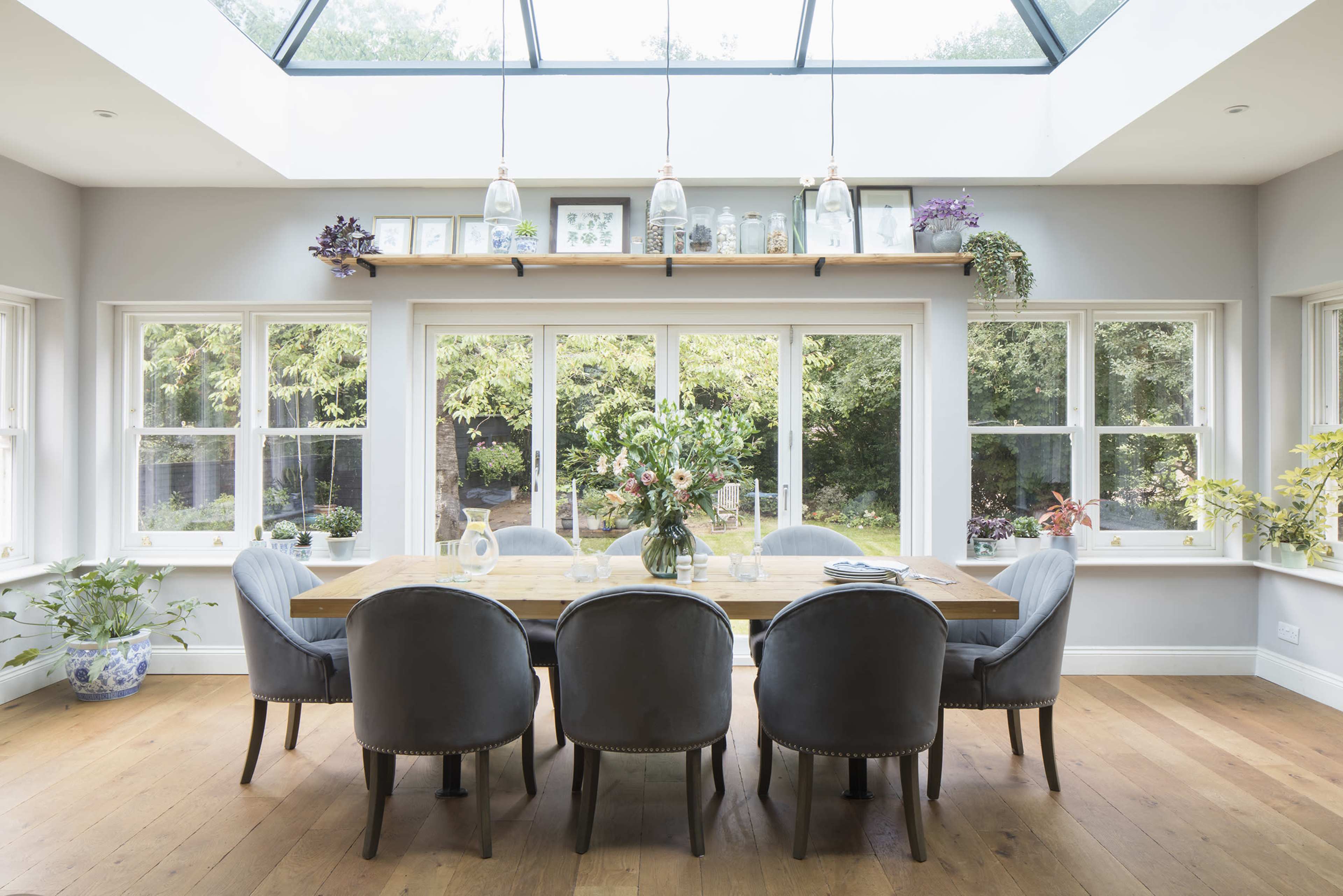 The dining room features a long wooden table surrounded by upholstered chairs, with large windows providing a view of the garden outside.