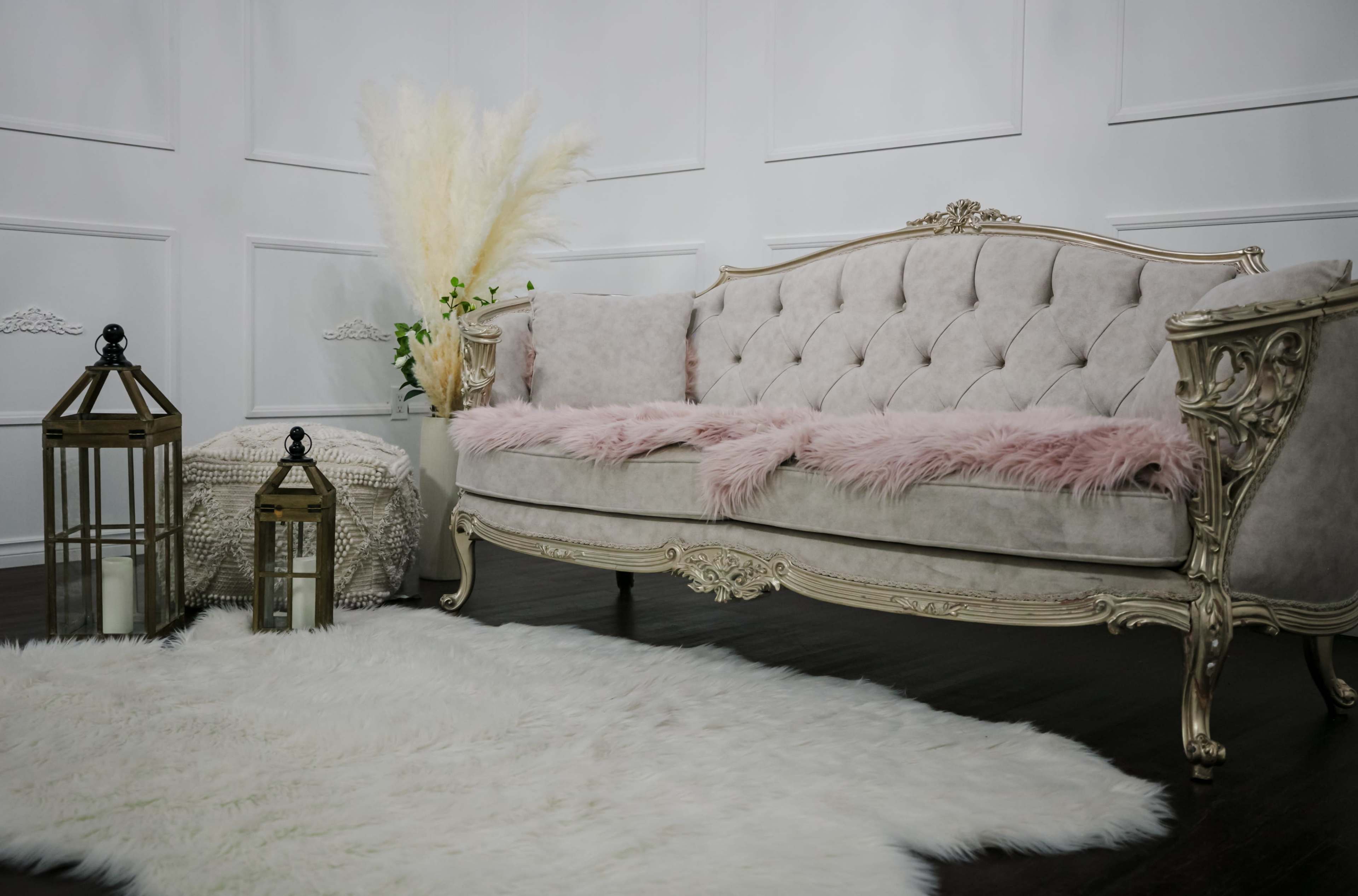 The scene features an elegant, upholstered sofa with decorative pillows, a textured side table, and two lanterns, all arranged on a fluffy white rug against a wall with paneling.