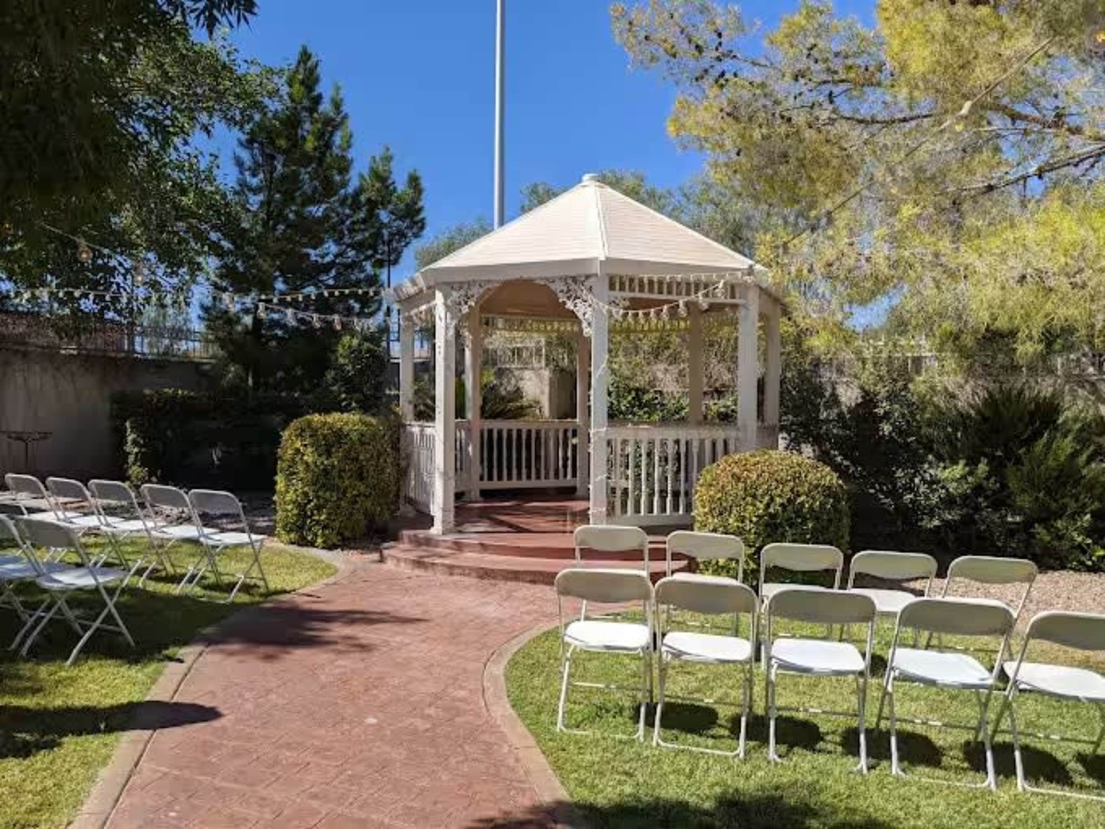 A white gazebo is positioned at the end of a brick pathway, surrounded by neatly trimmed landscaping and rows of folding chairs set up for an event.