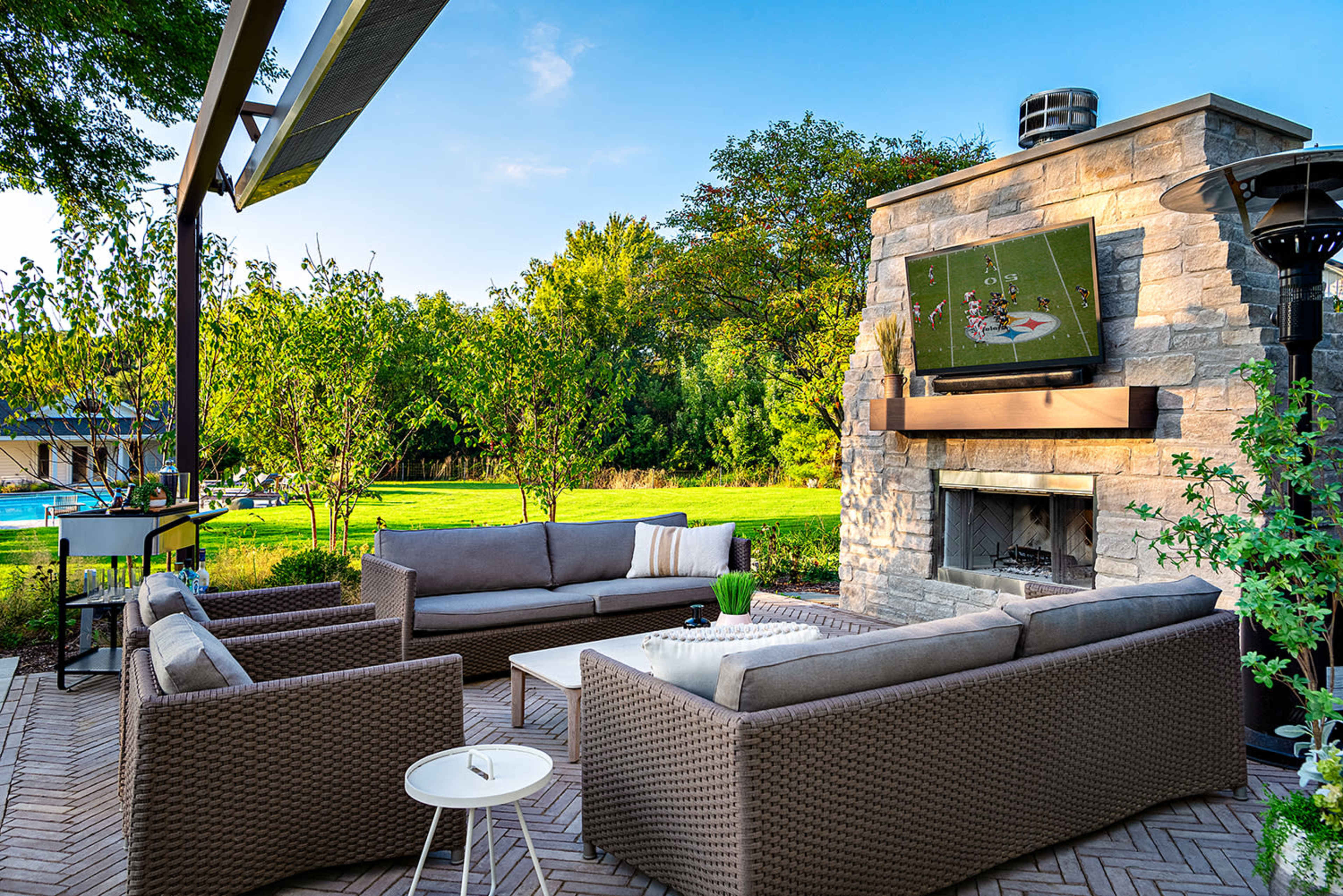 A patio with a seating area featuring wicker furniture and a mounted TV above a stone fireplace, surrounded by greenery and a view of a pool.