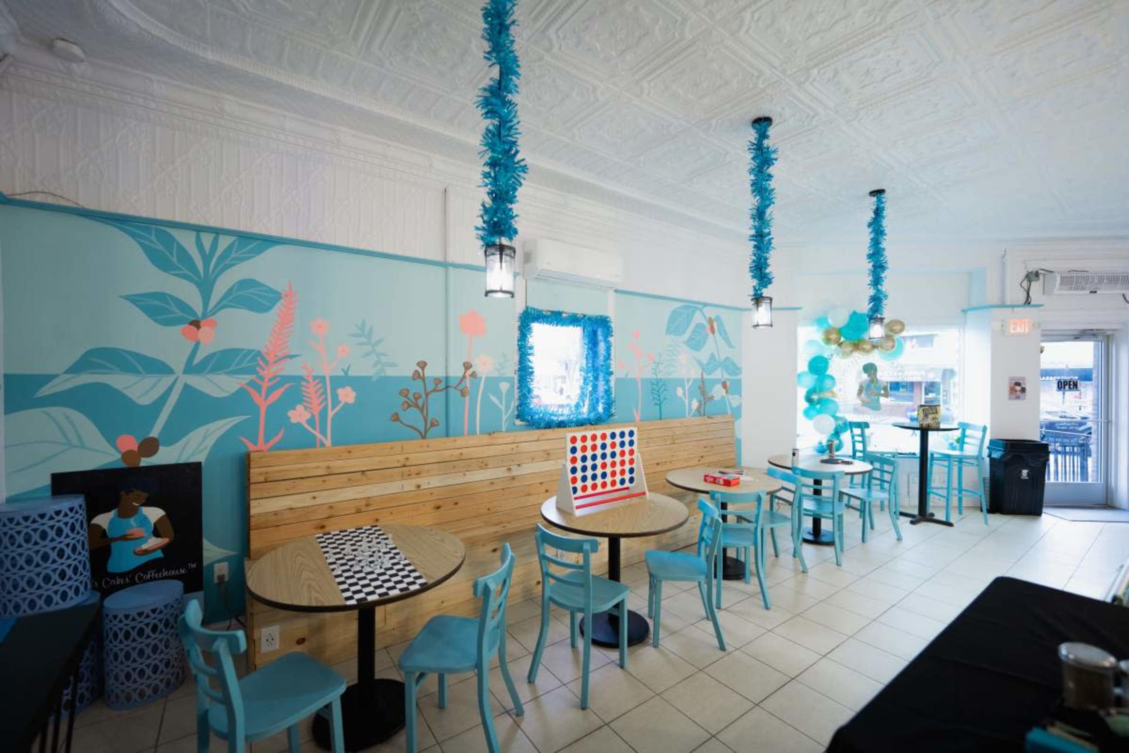 The image shows a bright, modern café interior with turquoise accents, wooden tables, and playful decorations.