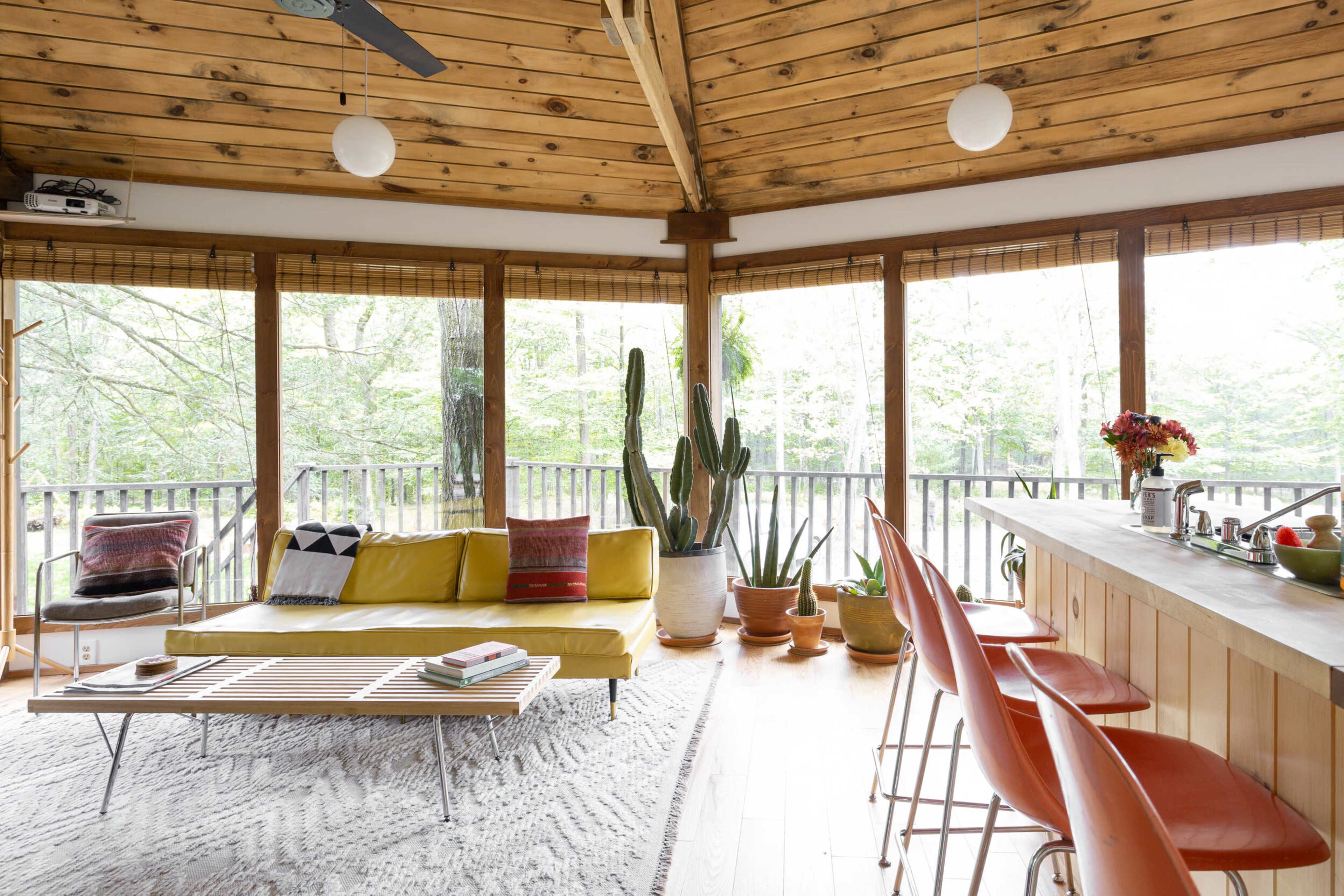 A modern, circular living space features a yellow sofa, a wooden ceiling, a bar with colorful stools, and large windows overlooking a forested area.