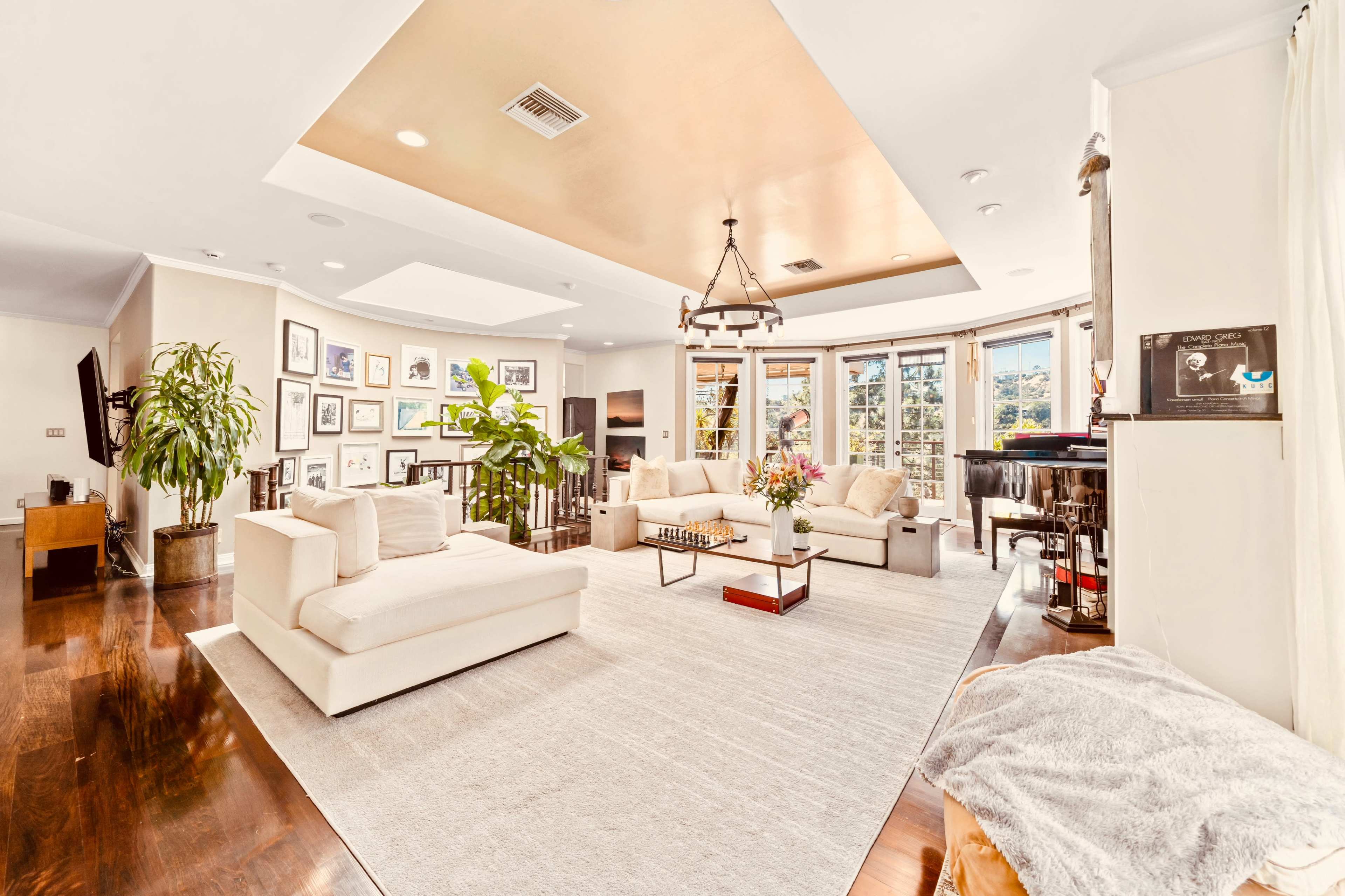 The image shows a spacious living room with a circular seating area, large windows, and a mix of furniture including sofas, a coffee table, and a piano.