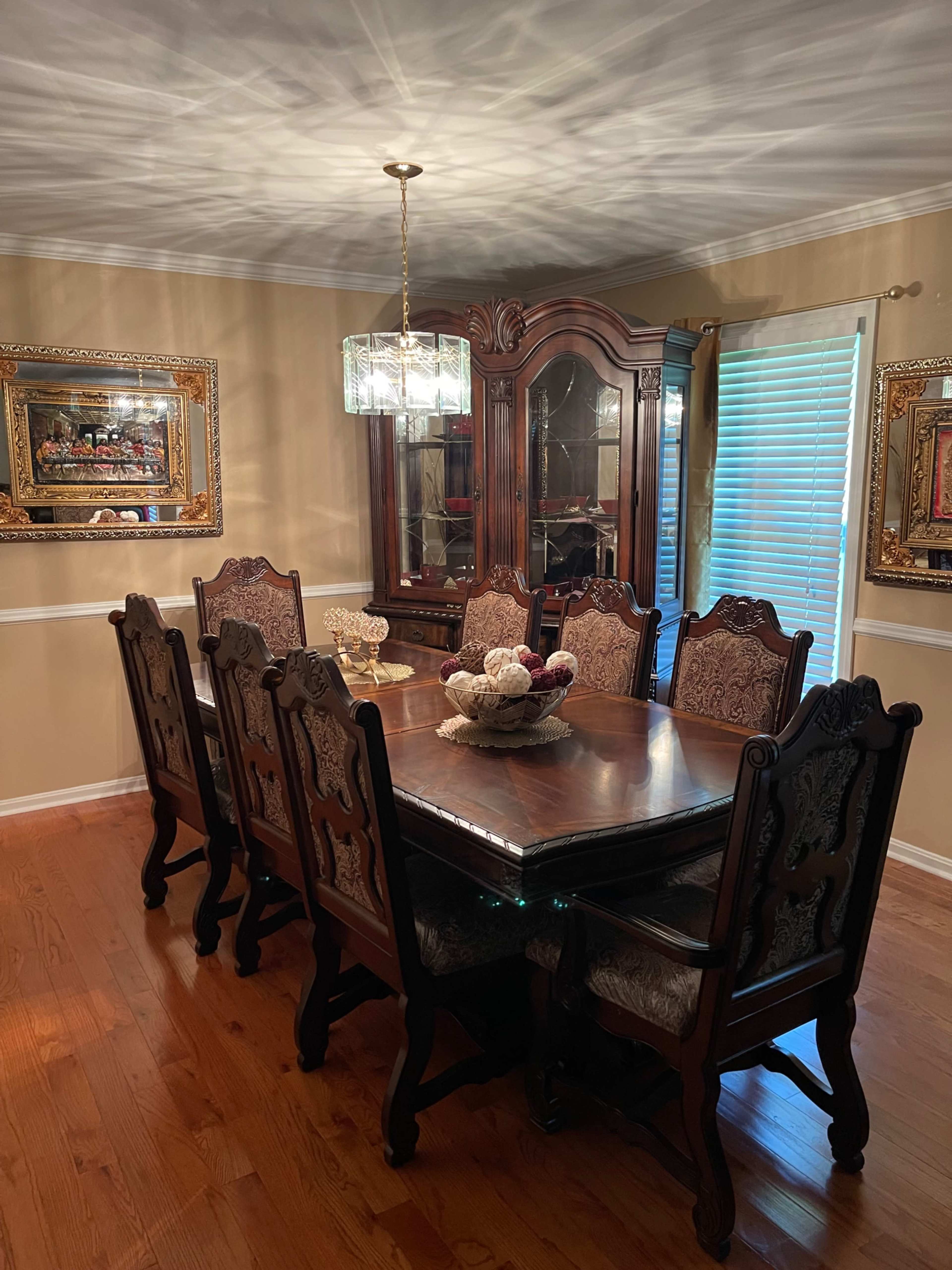 A wooden dining table with eight ornate chairs is centered in a warmly lit dining room, surrounded by elegant decor and a china cabinet.