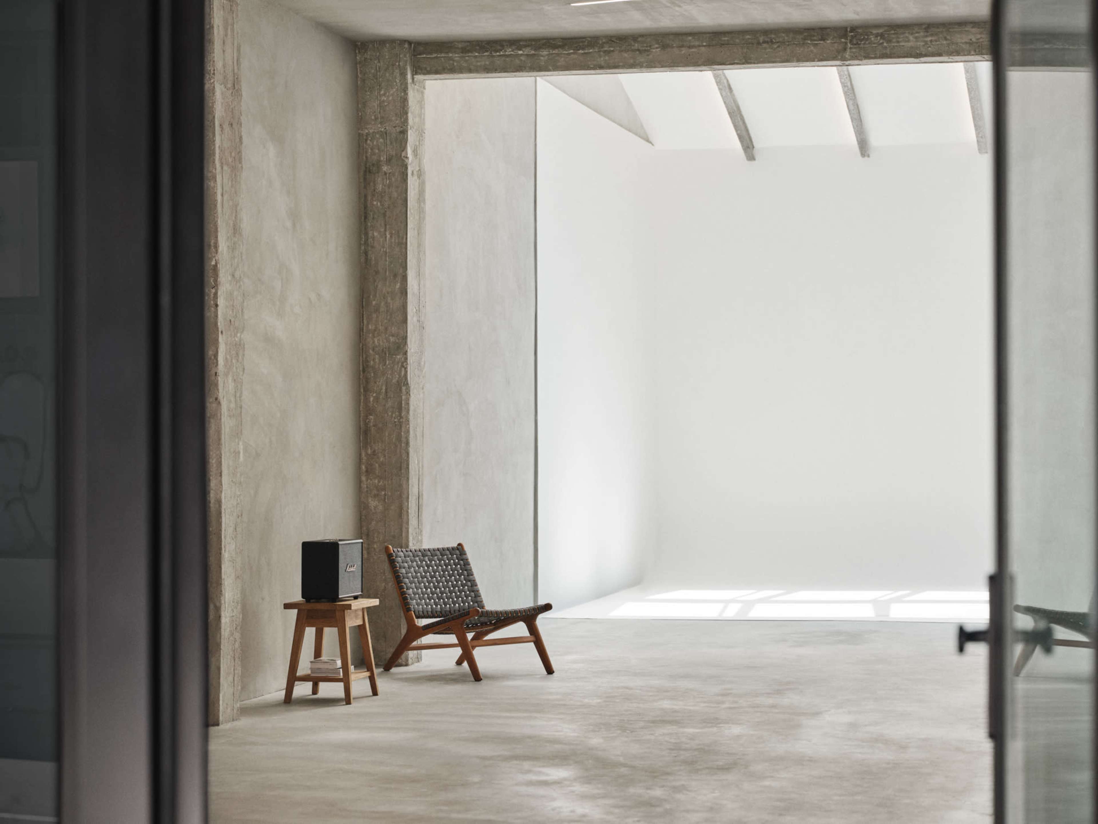 The image features a minimalist interior space with a wooden chair, a small speaker on a stool, and a large white wall.