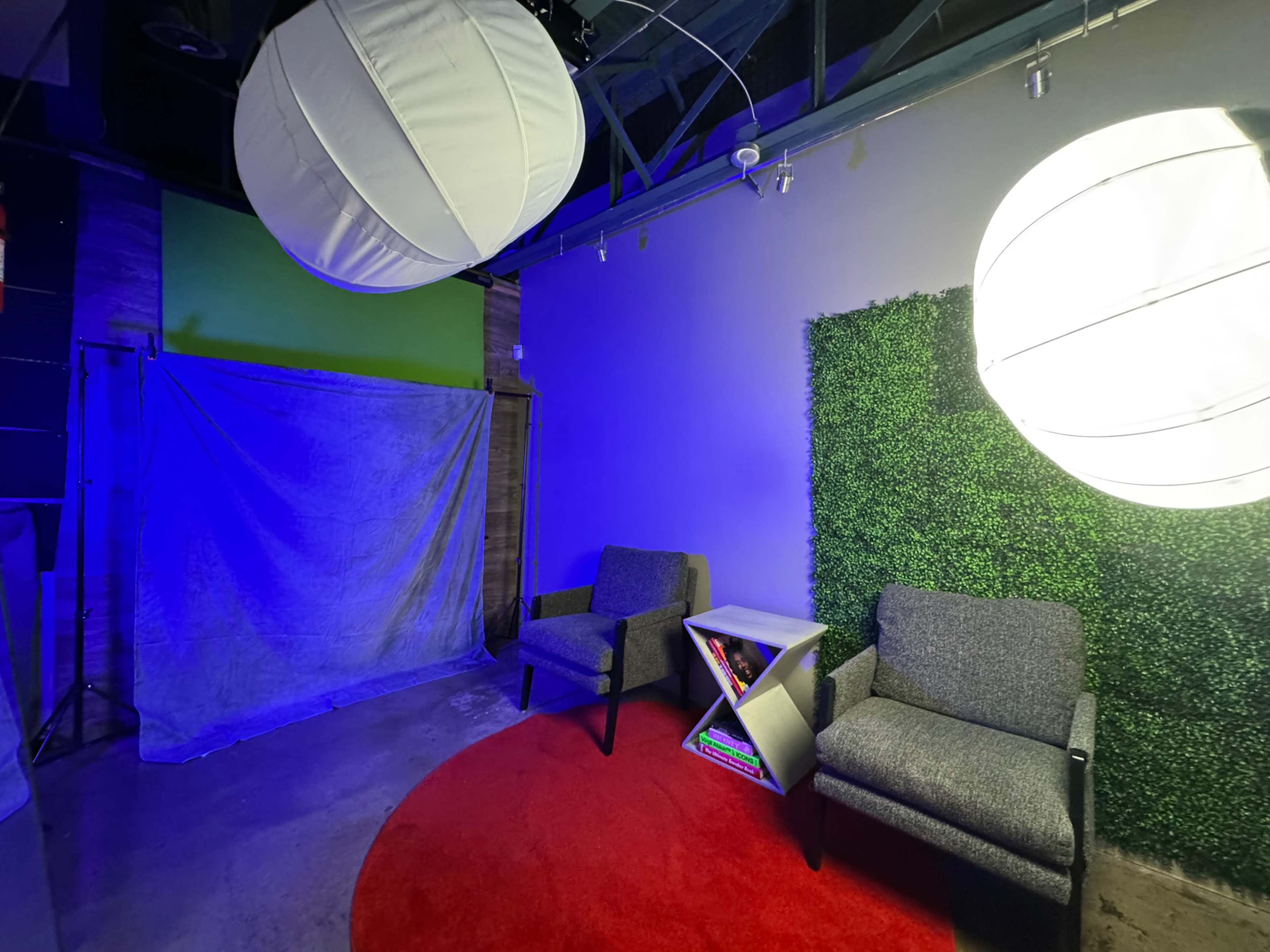 The image shows a small studio space with two large spherical lights overhead, a green backdrop, two gray chairs, and a small white side table on a red circular rug.