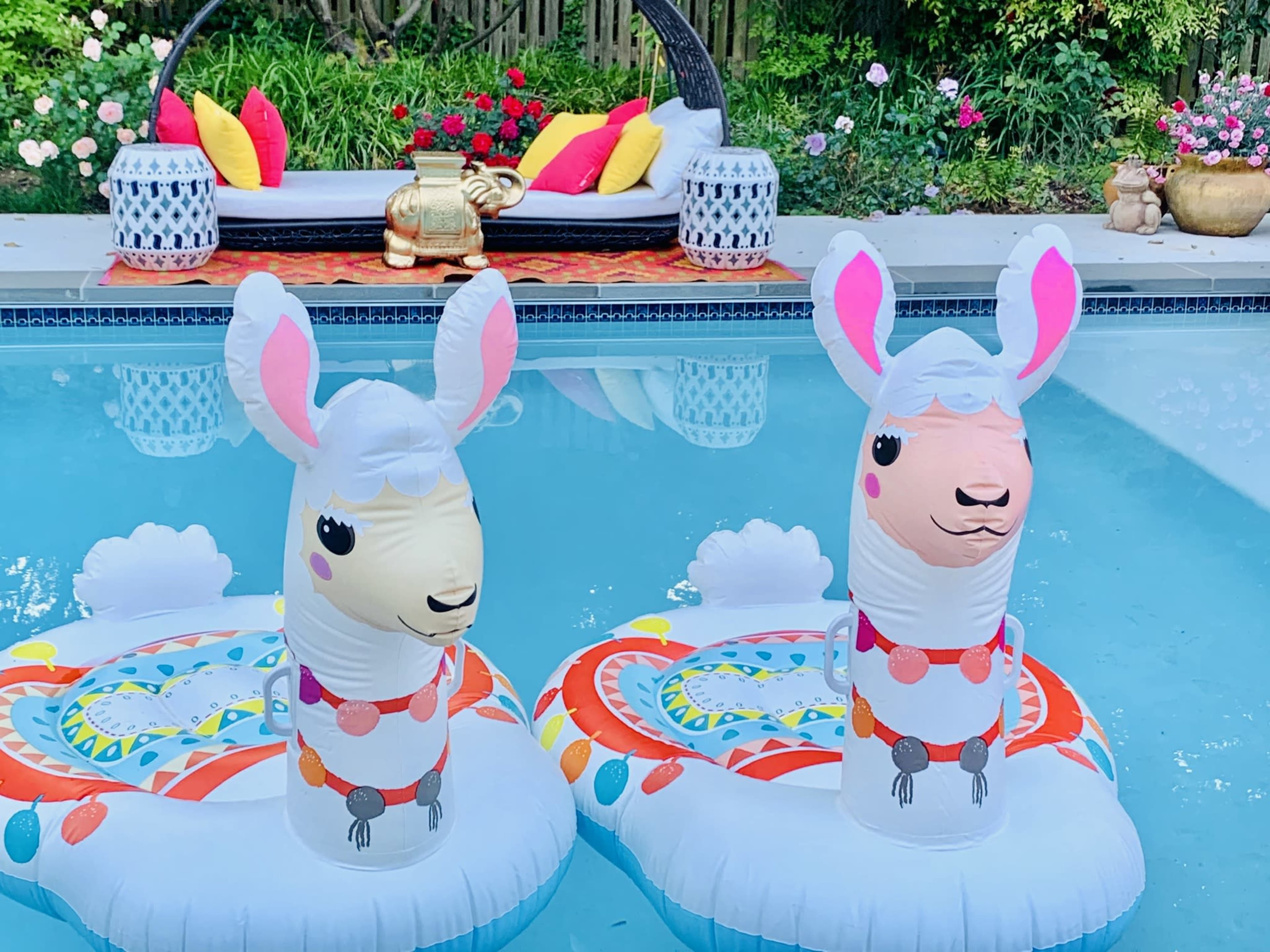Two inflatable llama pool floats are positioned in a swimming pool, surrounded by a landscaped yard featuring colorful flowers and a lounge area in the background.