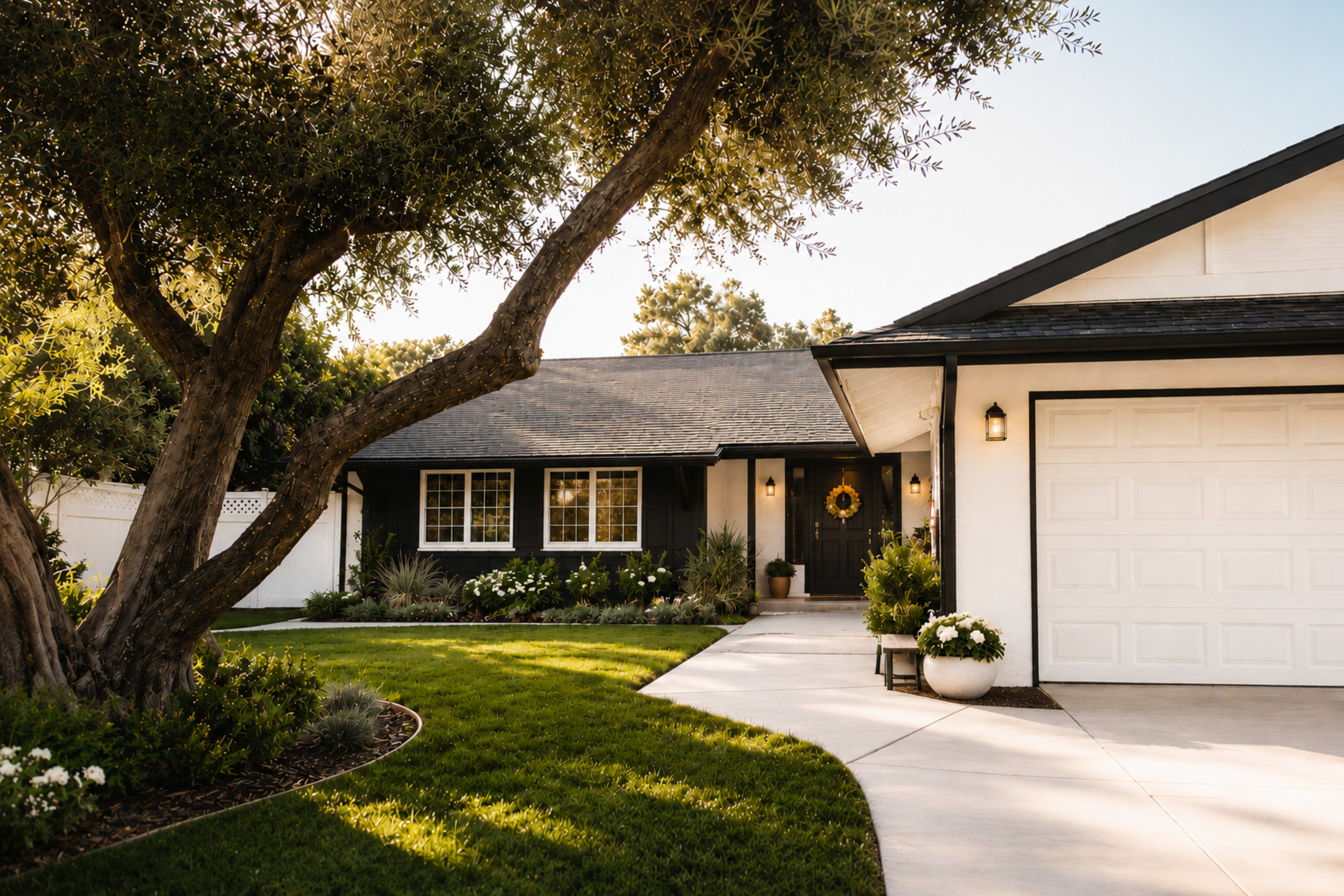 A modern single-story house with a gray roof, white garage, and a well-manicured lawn is framed by a large tree in the front yard.