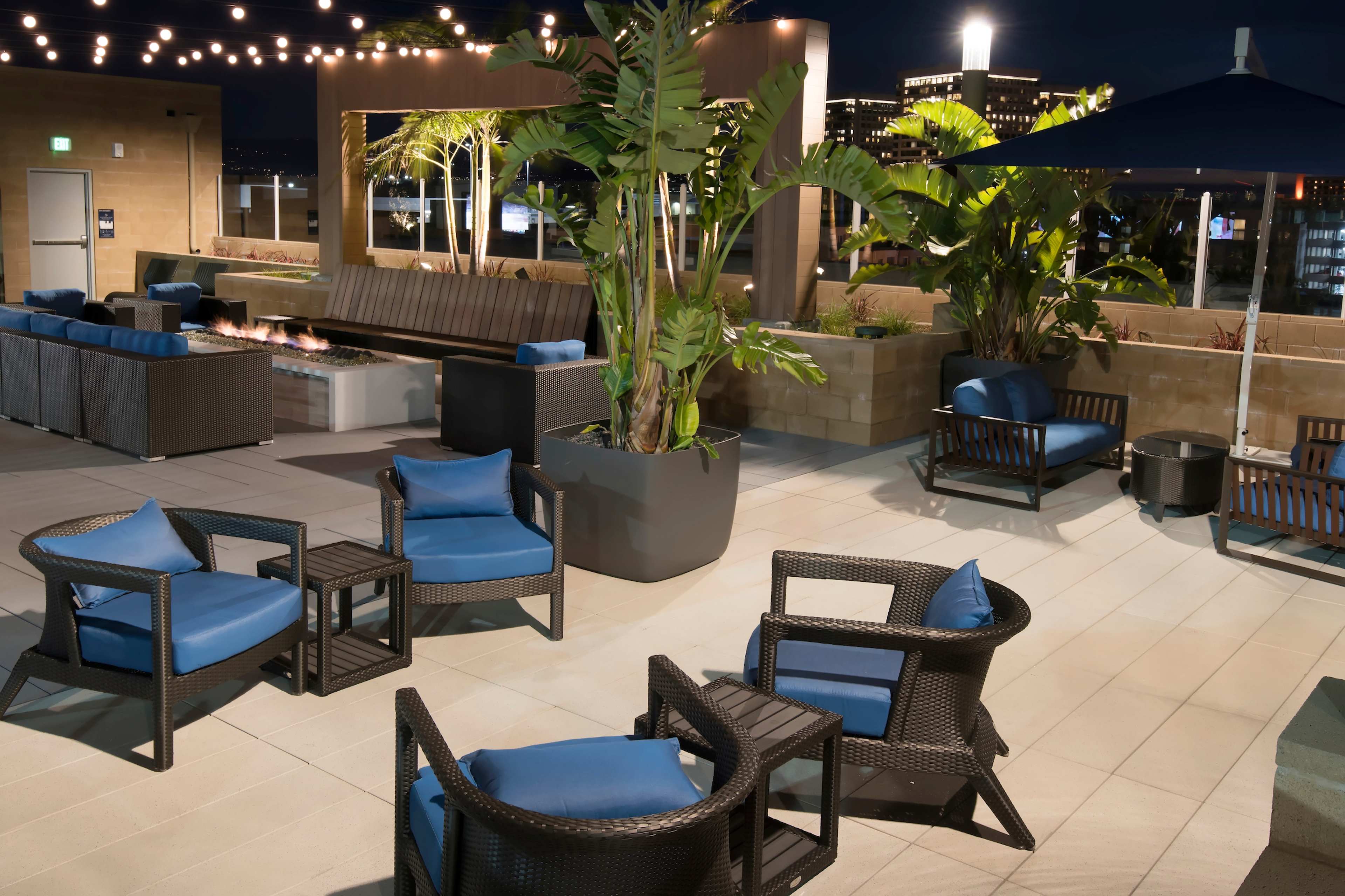 A rooftop lounge area with wicker furniture and potted palms, illuminated by string lights against a nighttime city backdrop.
