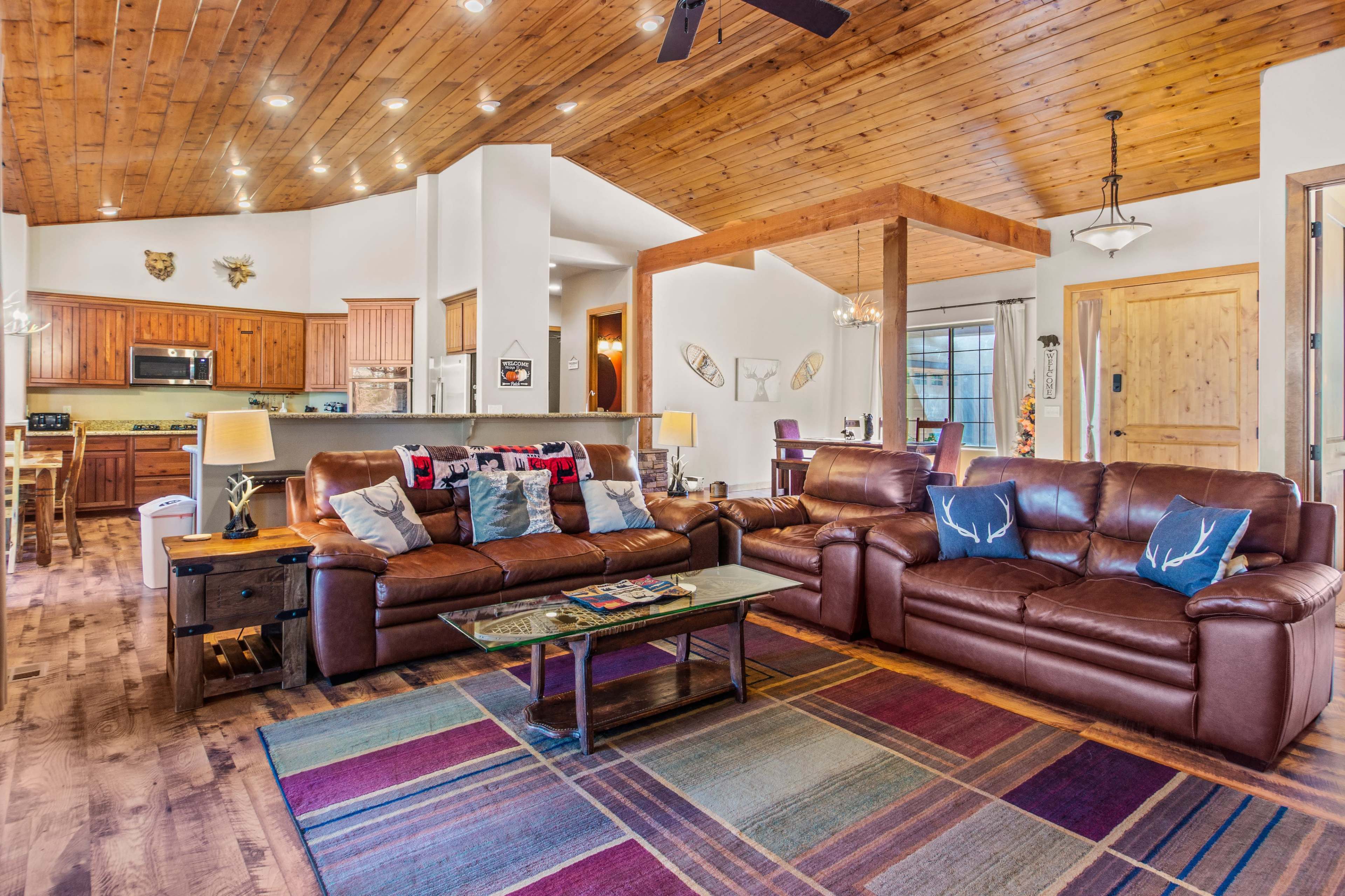 A spacious living room with two brown leather sofas, a coffee table, and a colorful area rug, surrounded by a kitchen and dining area.