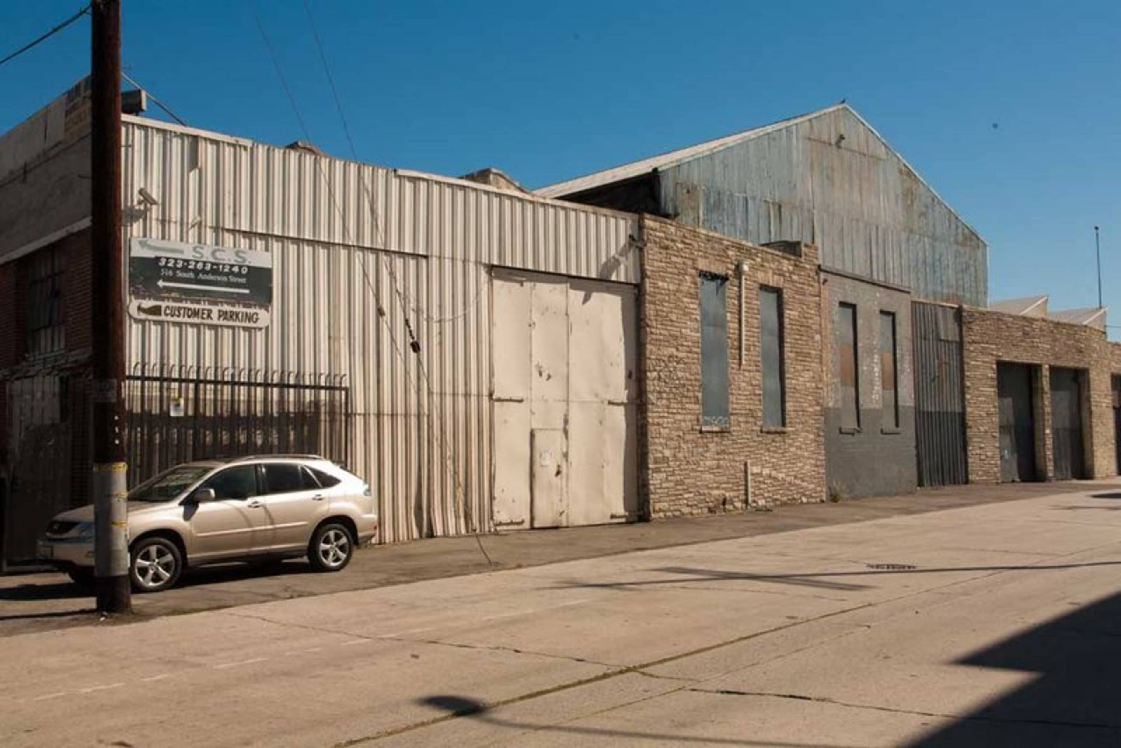 The image shows a narrow street lined with a tall, industrial building featuring metal siding and stone walls, alongside a parked silver car.