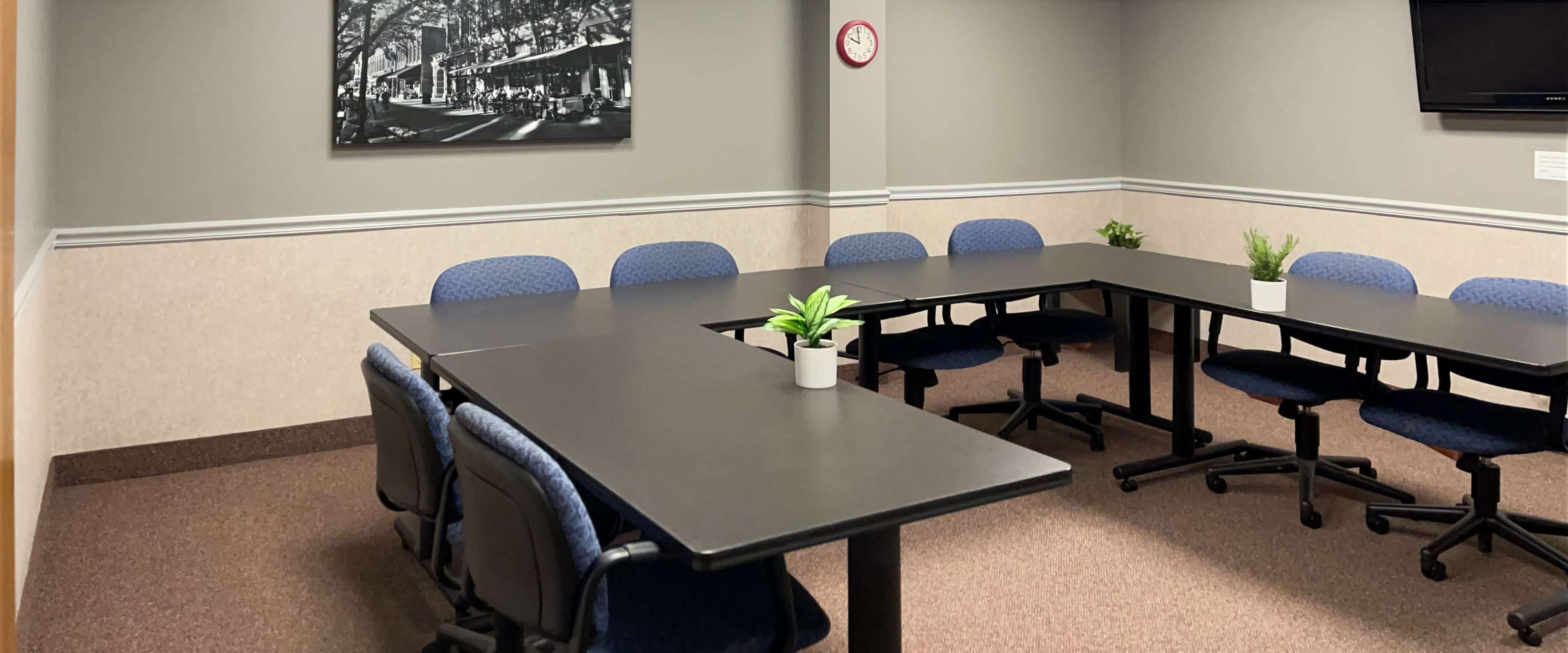 A conference room with two rectangular tables arranged in an L shape, surrounded by blue chairs and a small plant on the tables.