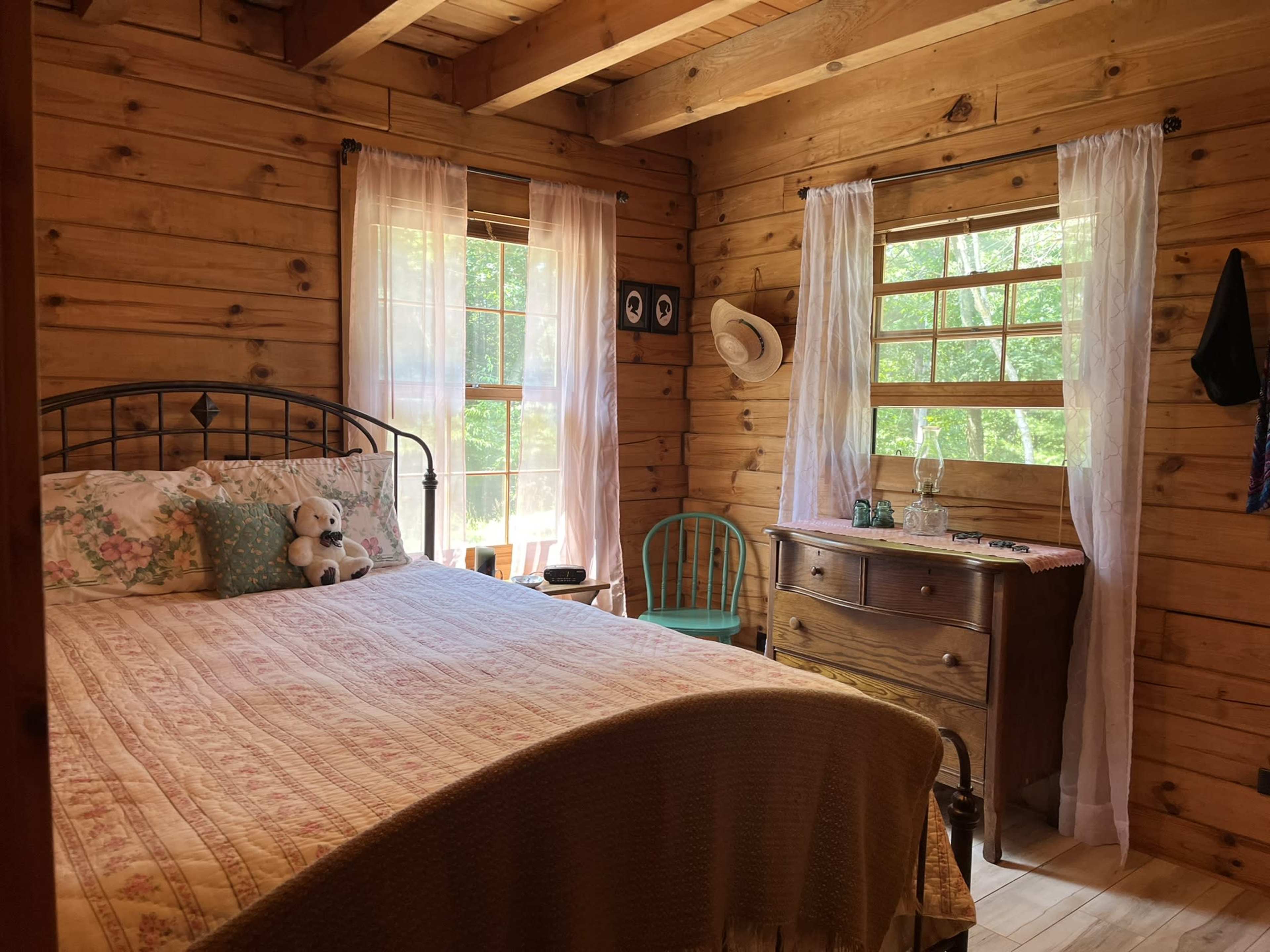 A cozy wooden bedroom with a bed, a dresser, and light streaming through the windows.