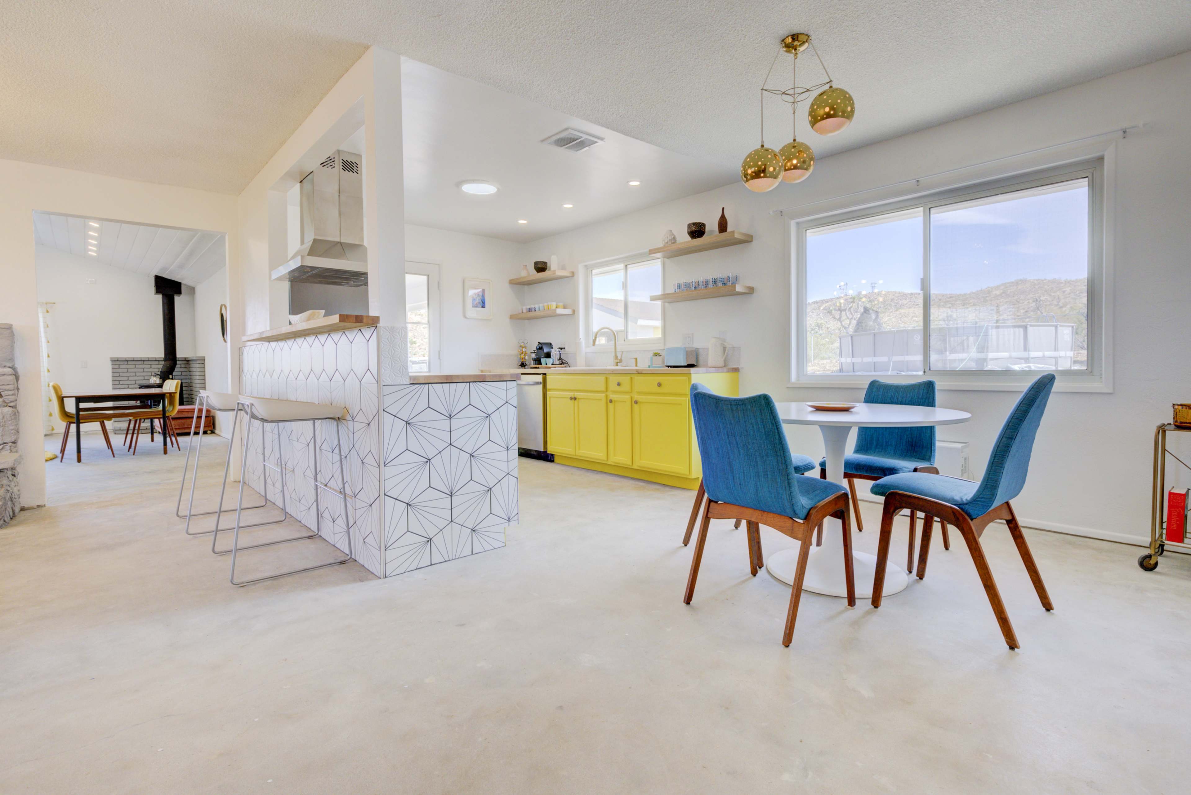 A modern kitchen and dining area features yellow cabinets, blue chairs, and a geometric-patterned bar counter, with large windows providing natural light.