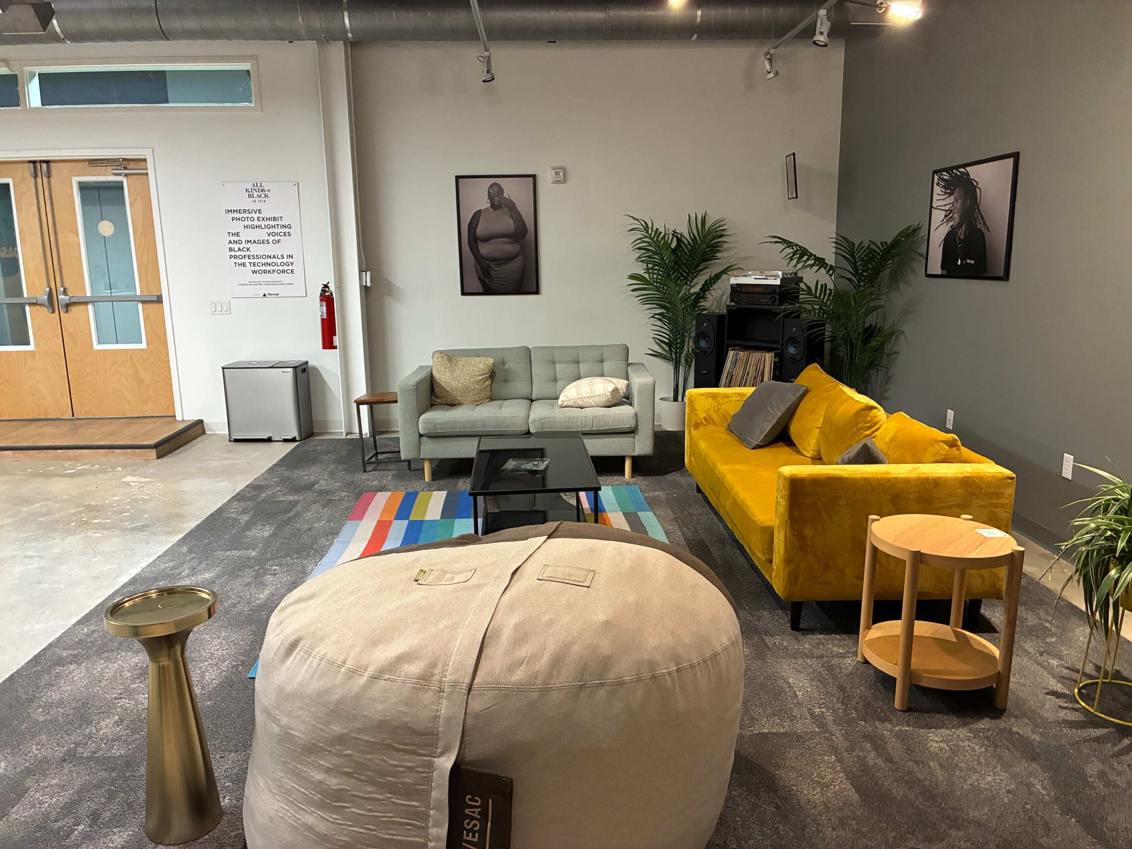 The image shows a modern lounge area with two sofas, a colorful rug, a large bean bag, a side table, and framed artwork on the walls.