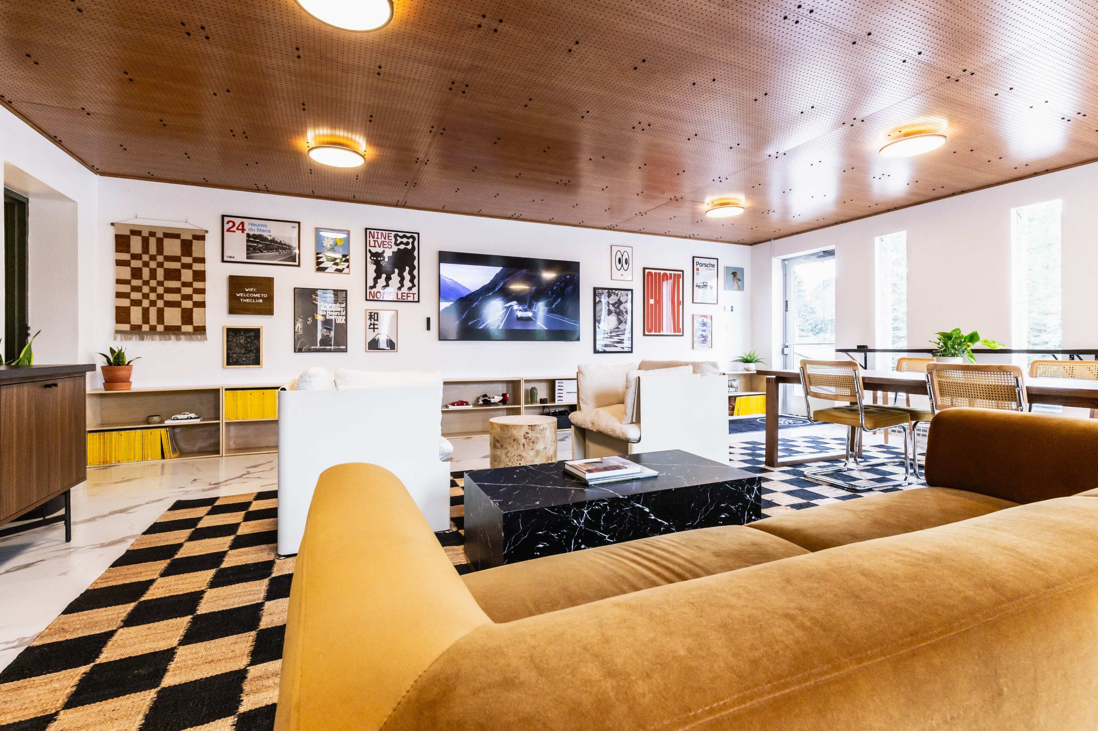 The image shows a modern living room featuring a television mounted on a wall, a coffee table, several framed artworks, and seating arrangements on a patterned rug.