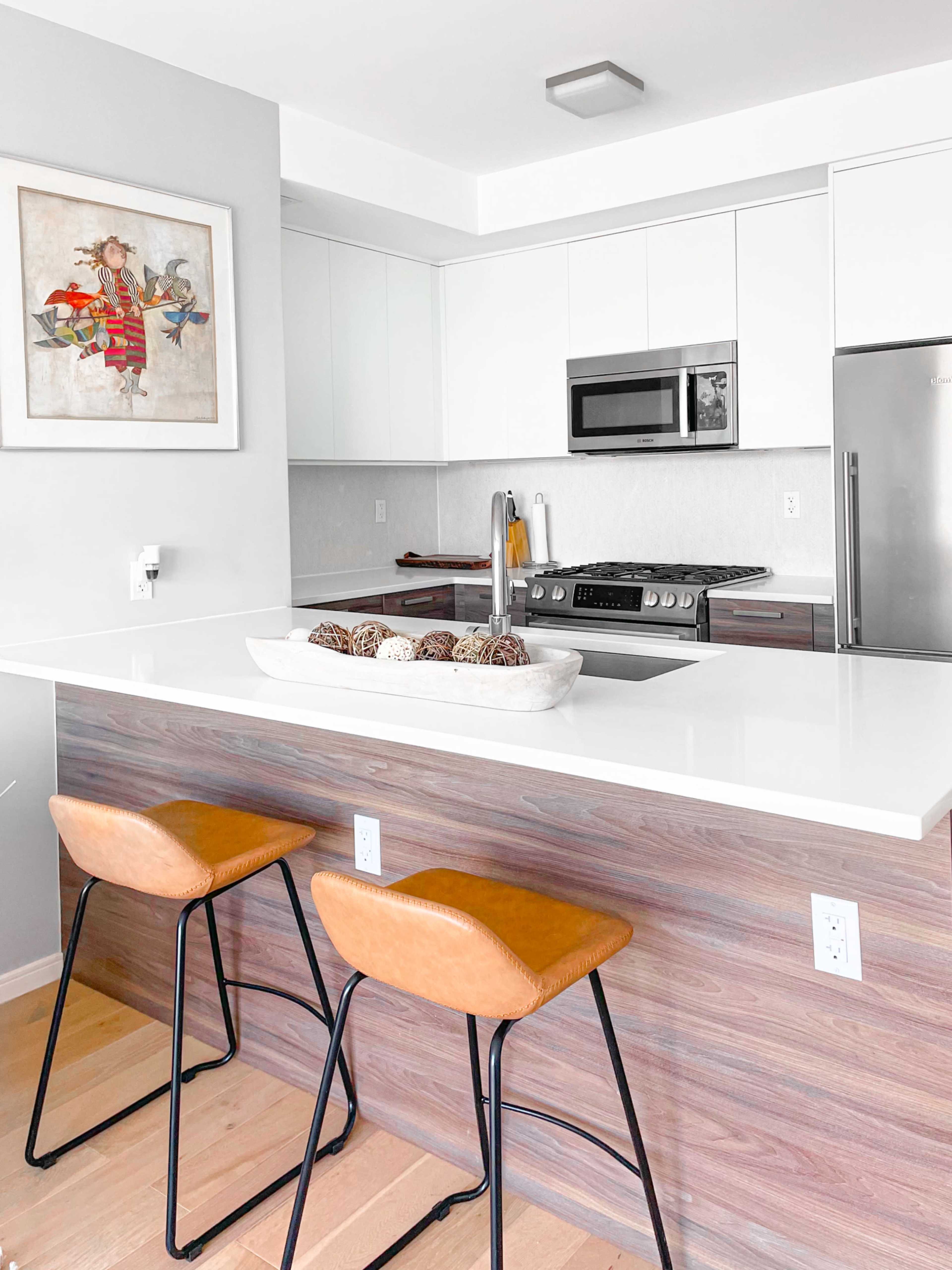 A modern kitchen features a central island with two leather-seated stools, stainless steel appliances, and a decorative bowl of natural elements.