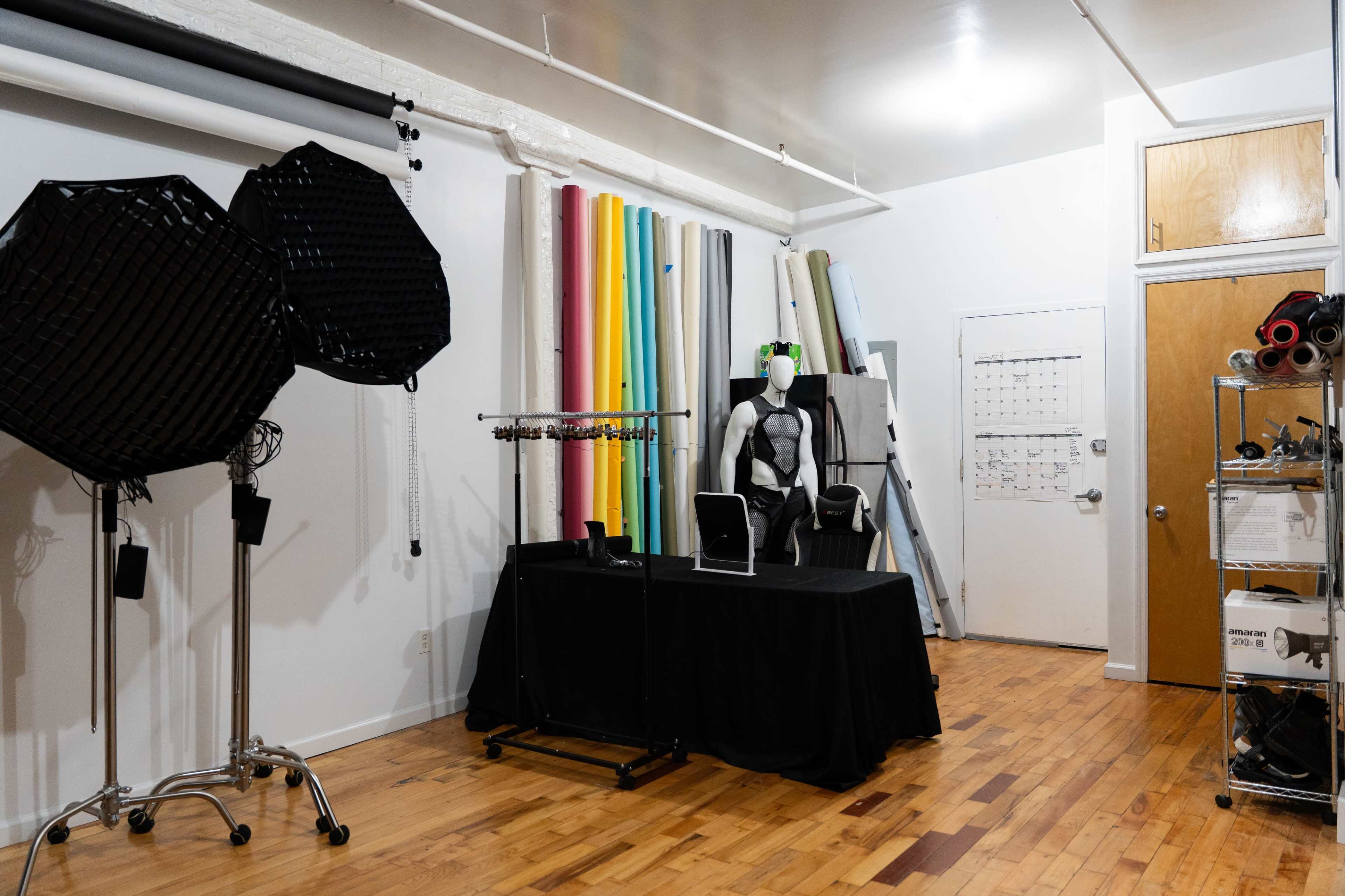 The image shows a photography studio with colorful backdrop rolls on the wall, a mannequin dressed in a costume, and various lighting equipment arranged on a black table.