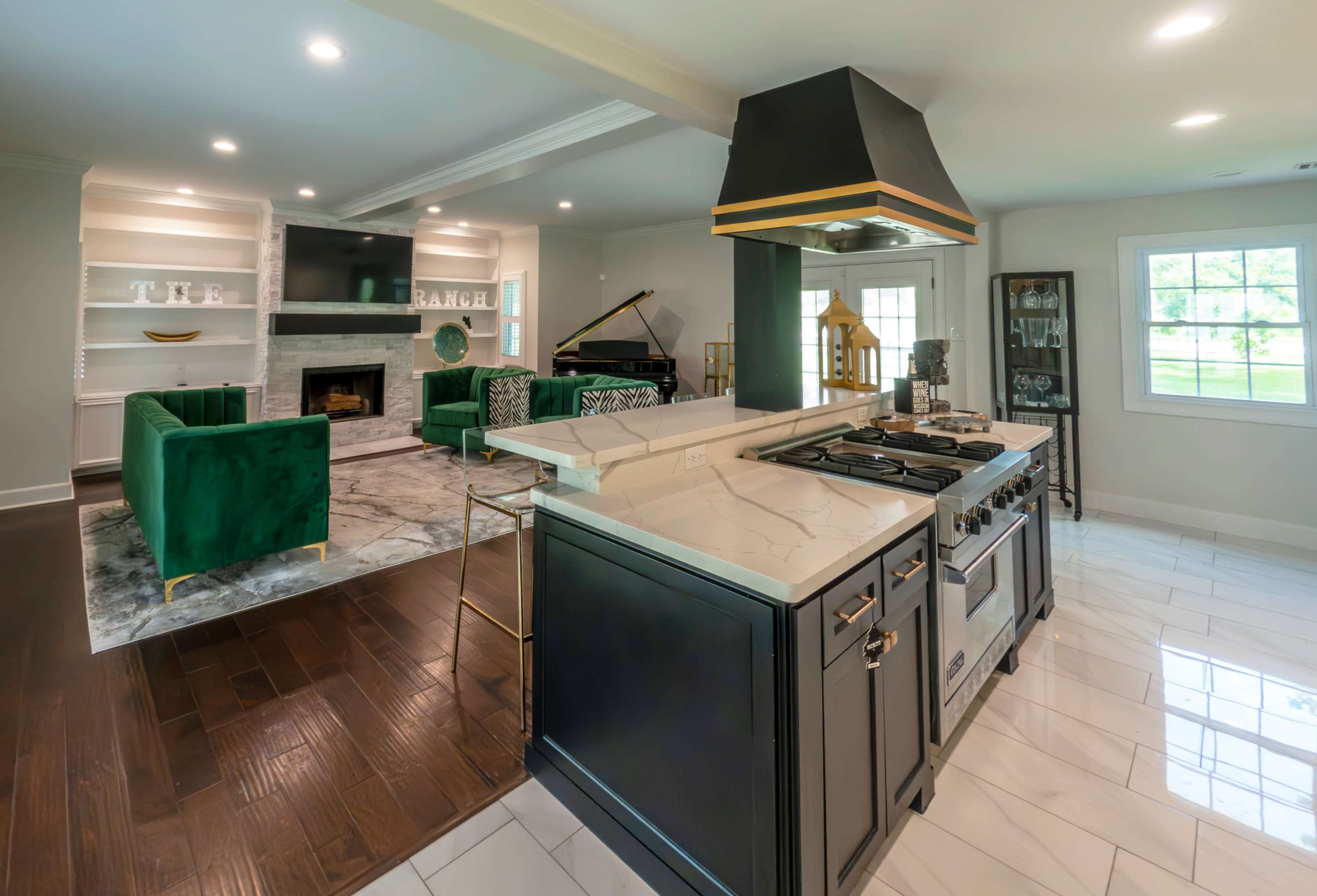 The kitchen features a central island with a gas stove and a large hood, opening to a living area with green upholstered furniture and a piano.
