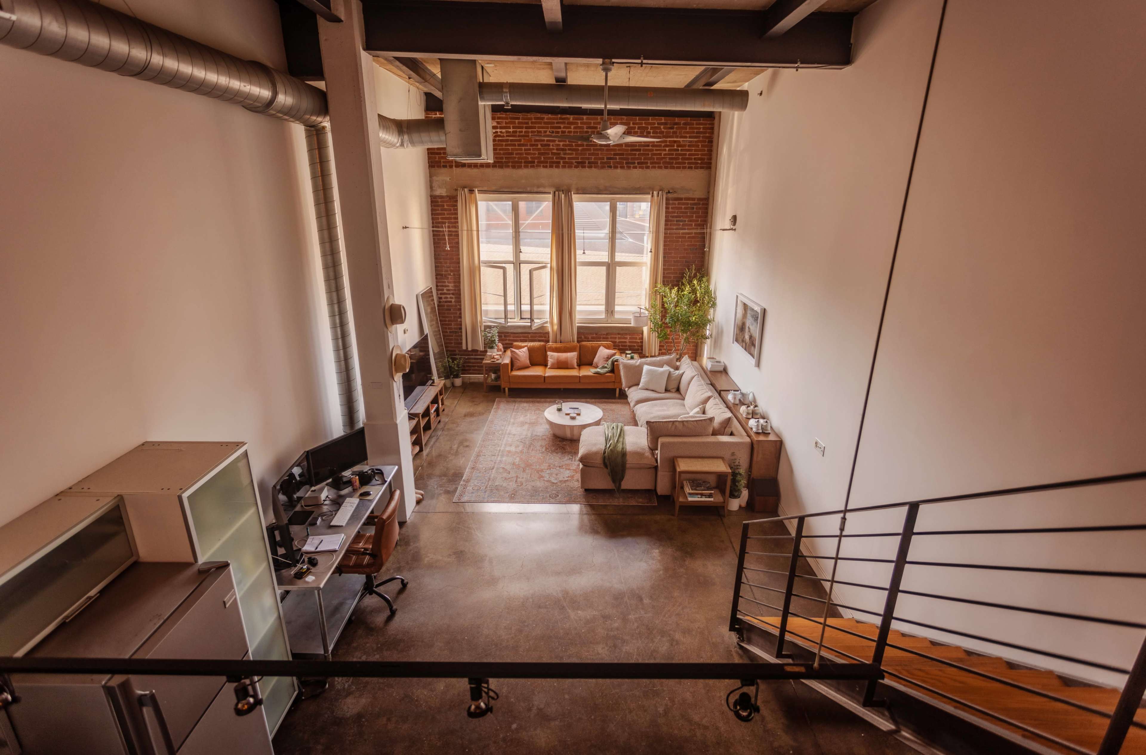The image shows a spacious, modern loft with large windows, hardwood flooring, and a living area furnished with a sofa and seating arrangement.