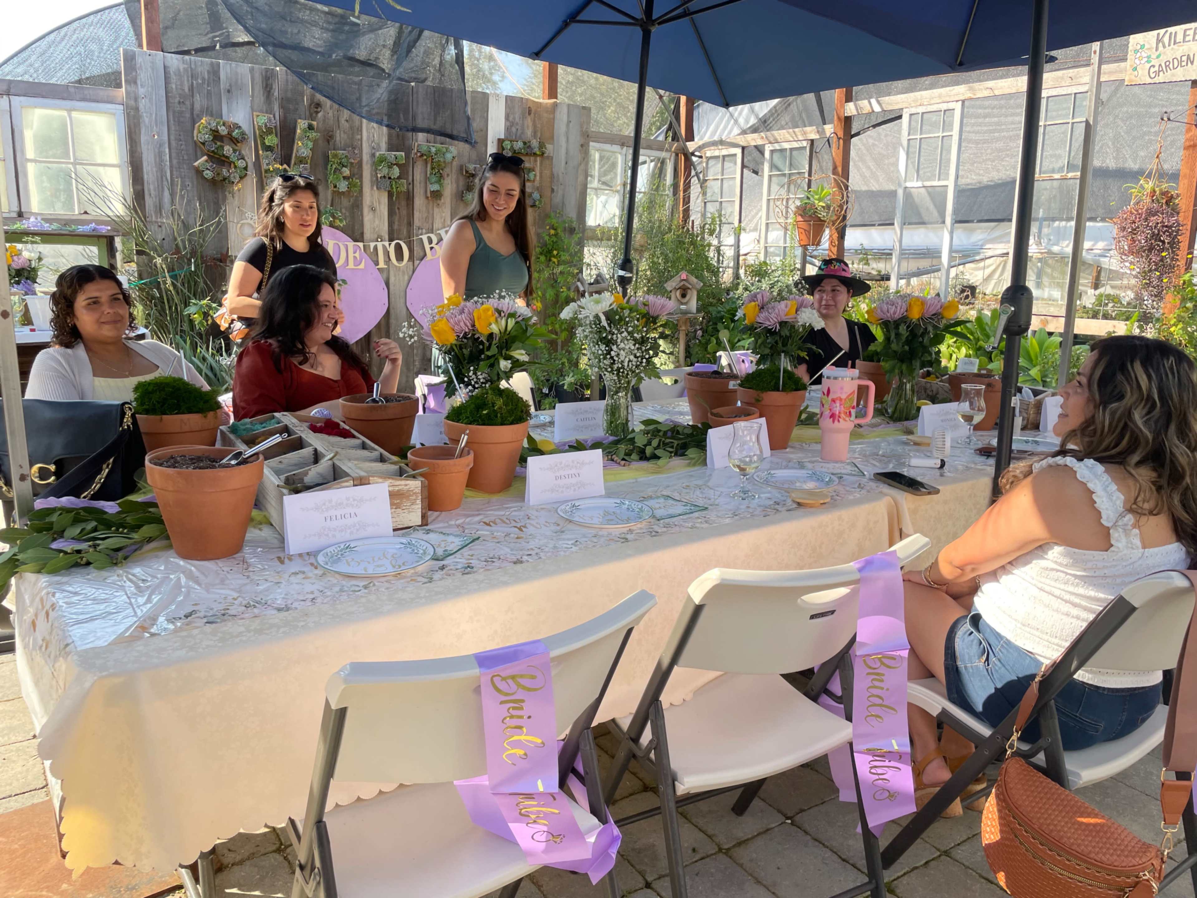 A group of women sits at a table decorated with potted plants and flowers in a greenhouse setting.