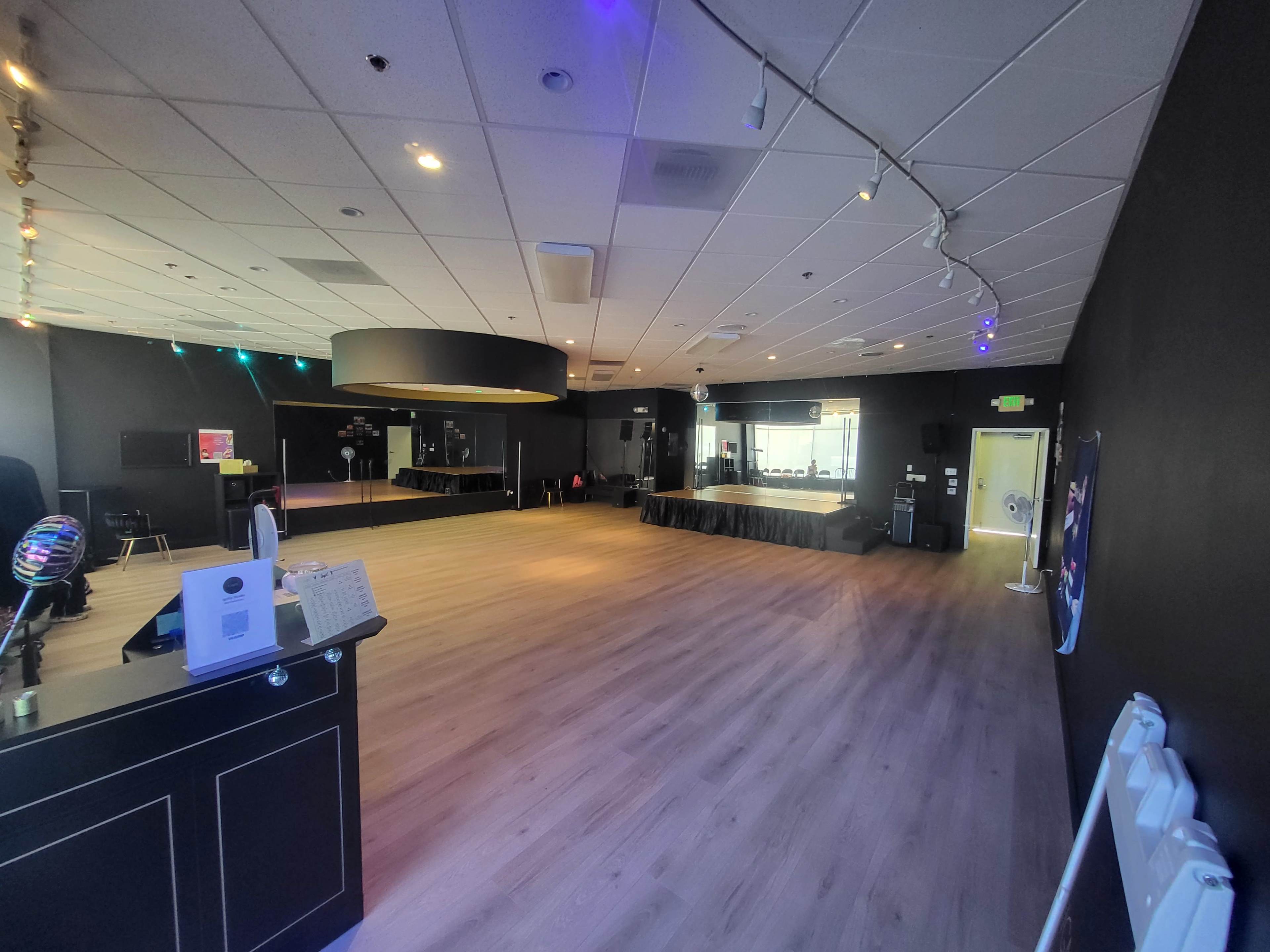 The image shows an empty event space with wooden flooring, black walls, and a stage area visible in the background.