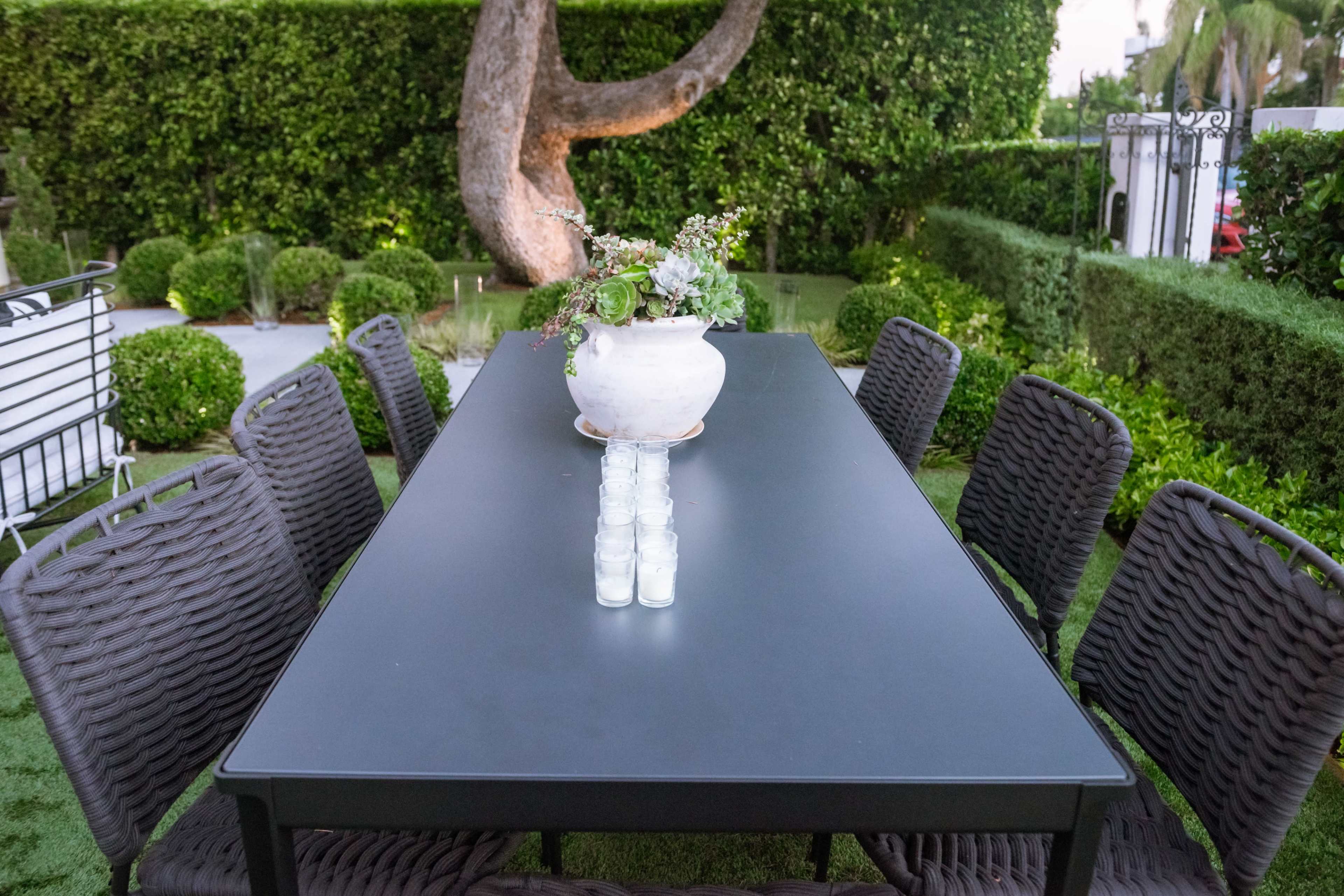 A long black dining table is set outdoors with a white vase of flowers in the center, surrounded by neatly arranged chairs and lush green hedges.